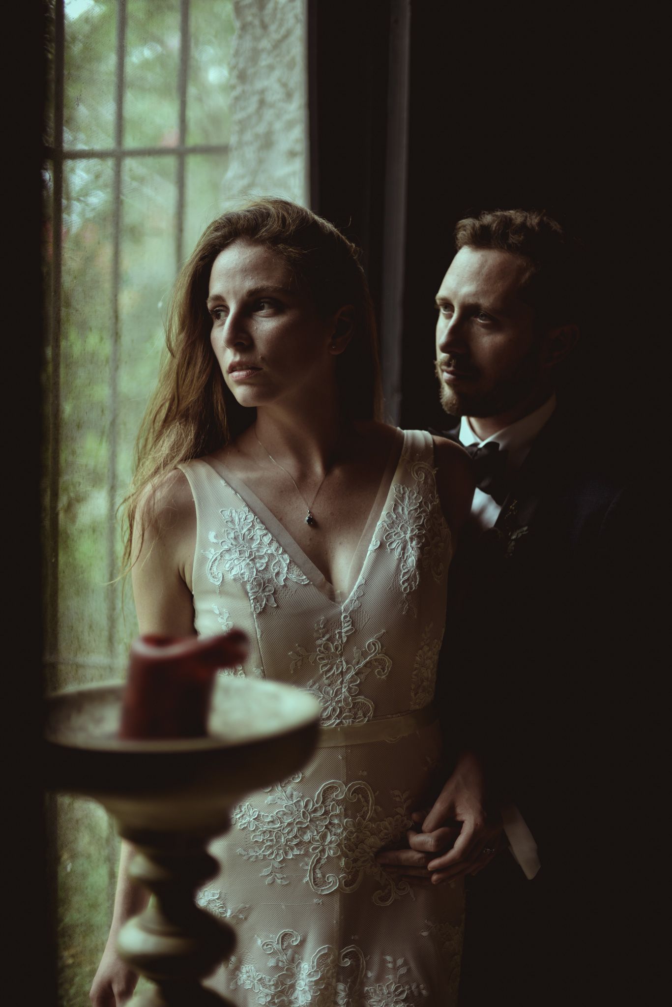 A moody wedding portrait in front of a window shows a couple in formal attire with dramatic lighting and vintage atmosphere.