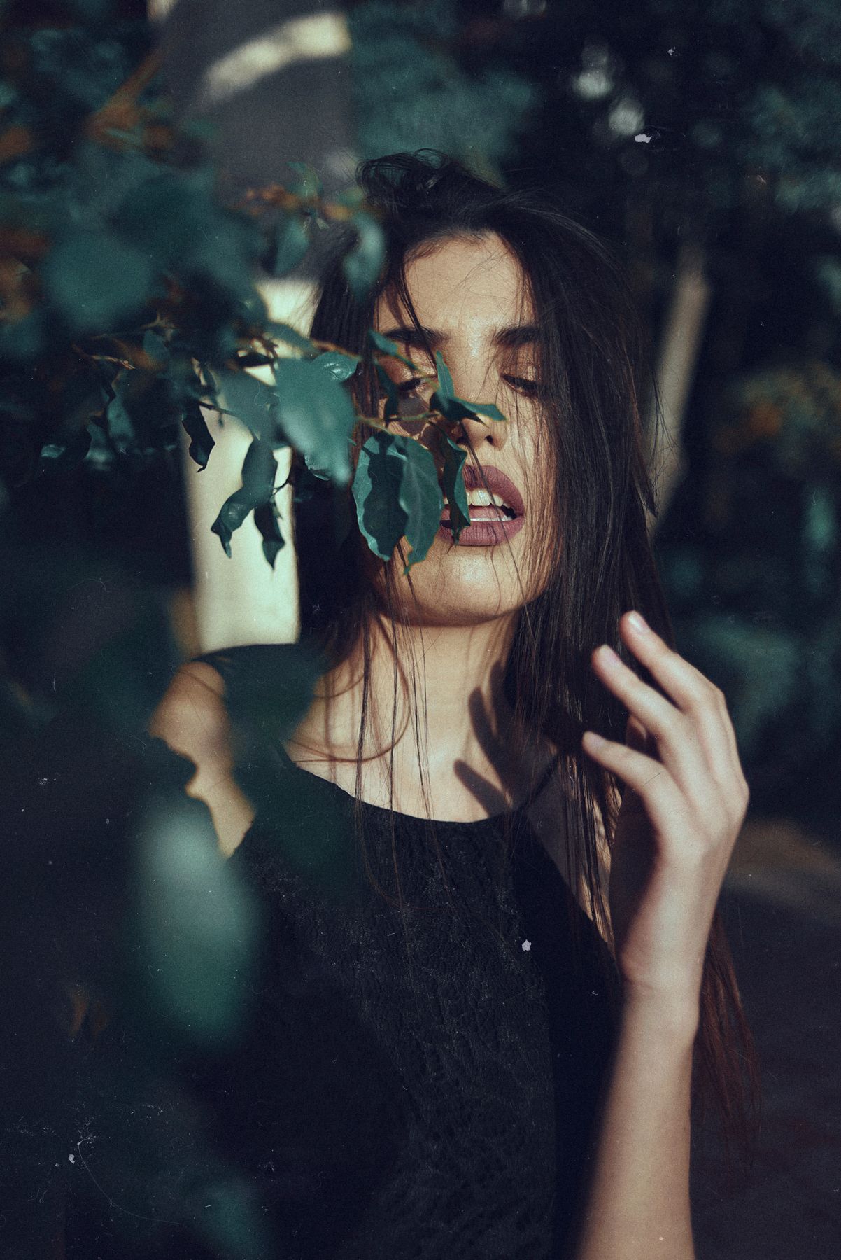 Moody portrait through leaves showing dark-haired figure in black against blurred background.