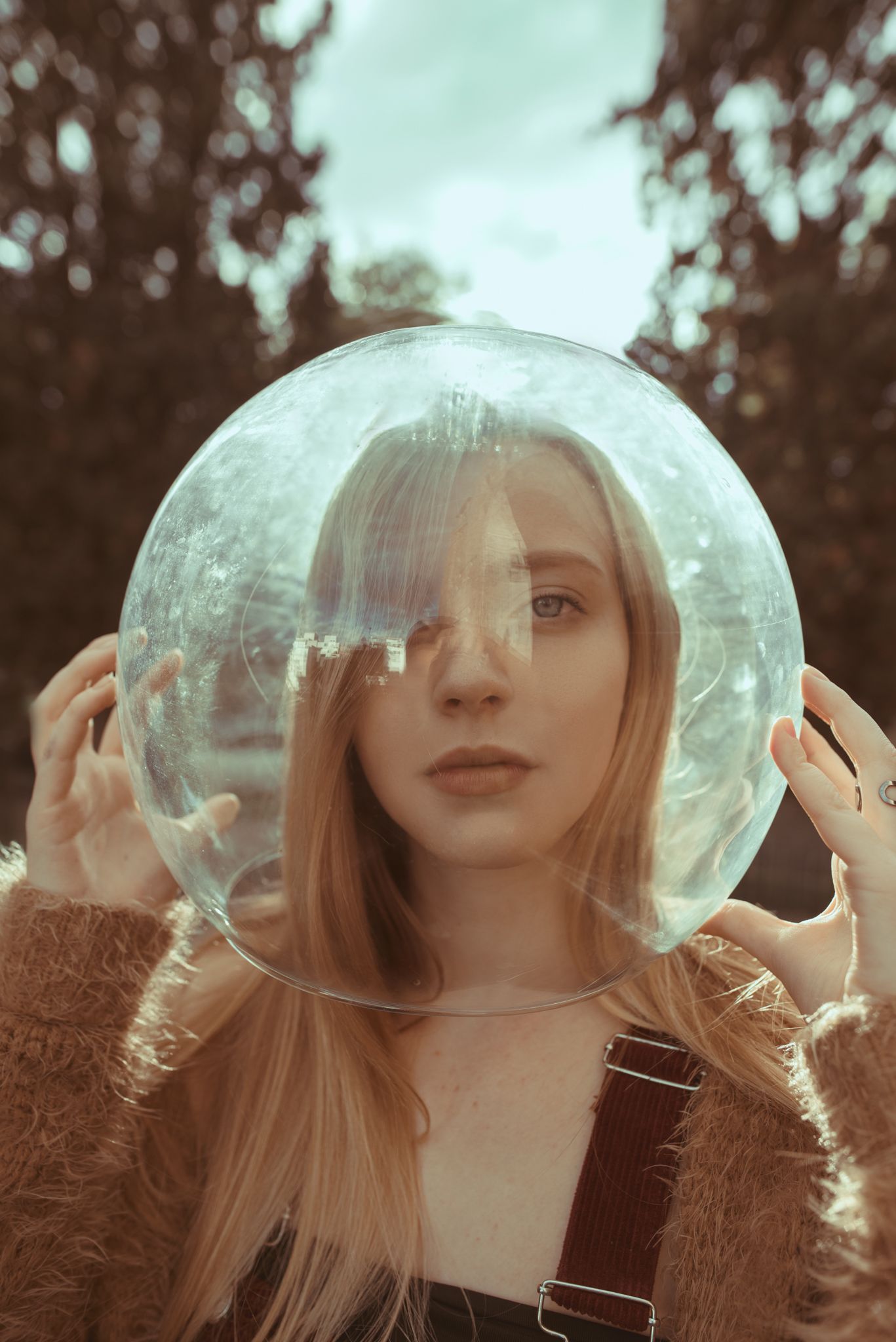 Woman wearing fuzzy sweater holds clear glass sphere in dreamy forest setting.