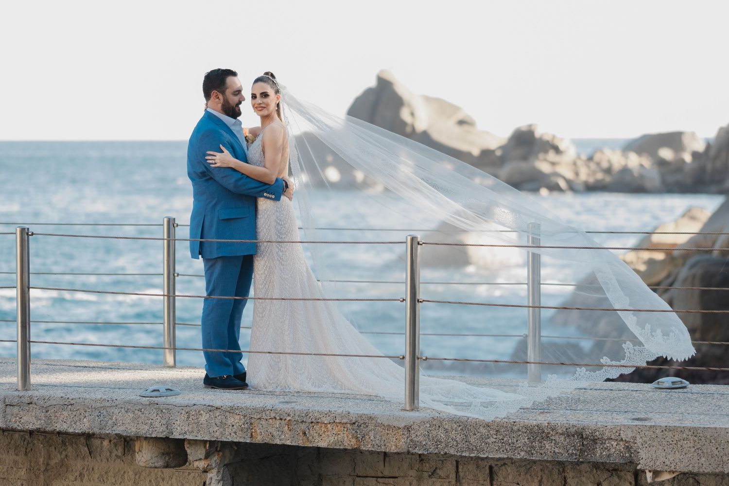 Couple shares romantic moment on oceanside pier as bride's veil flows in the wind.