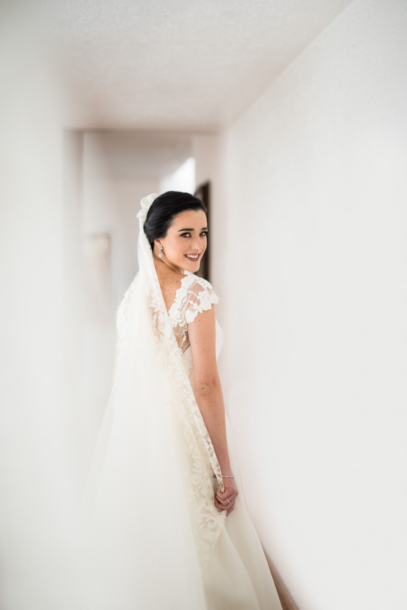Bride in lace gown poses elegantly in minimalist white hallway.