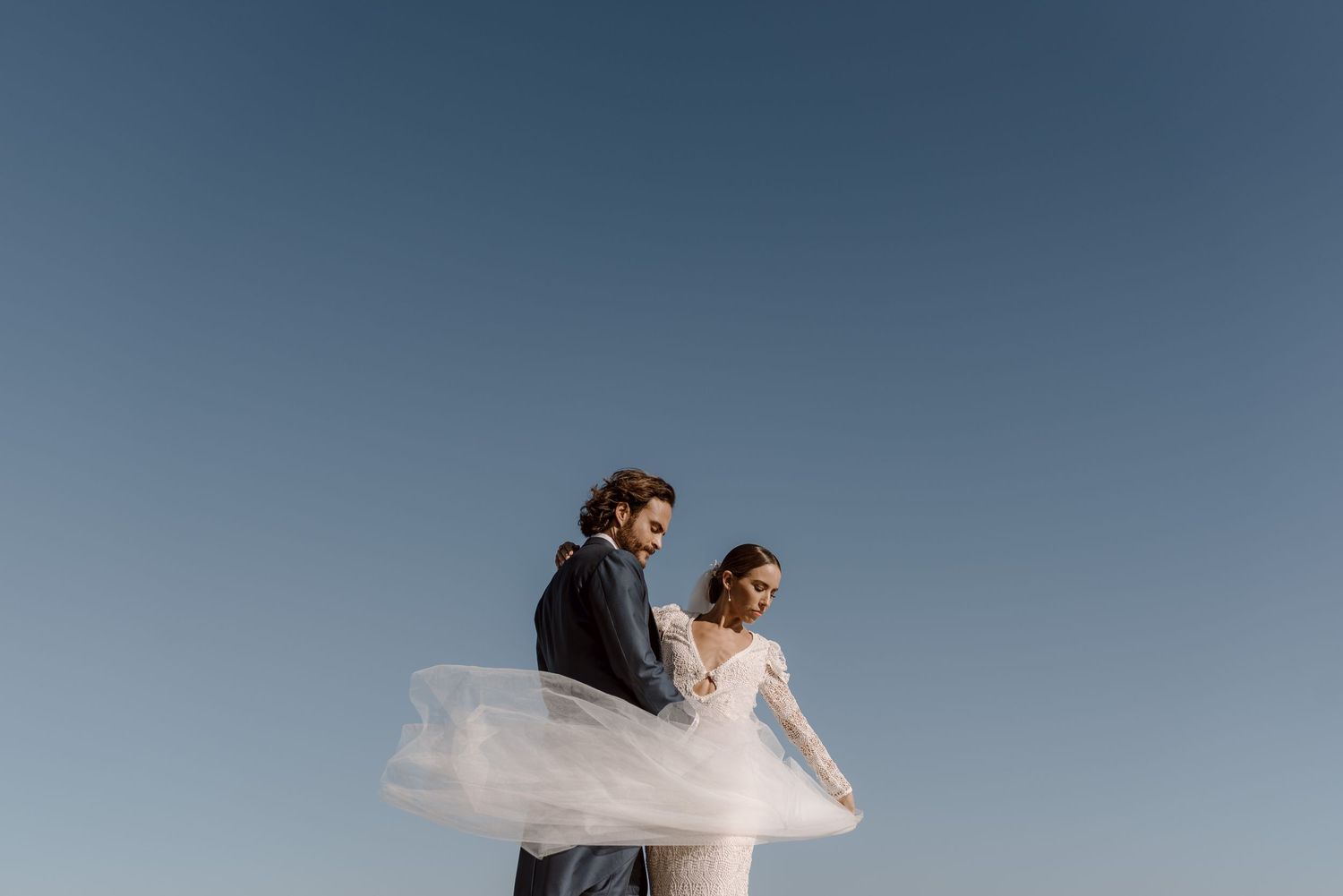 Ethereal wedding portrait with flowing dress against clear blue sky.
