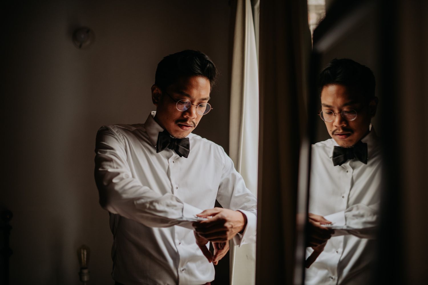 Person in white dress shirt and bow tie checking time while standing near window with reflection.