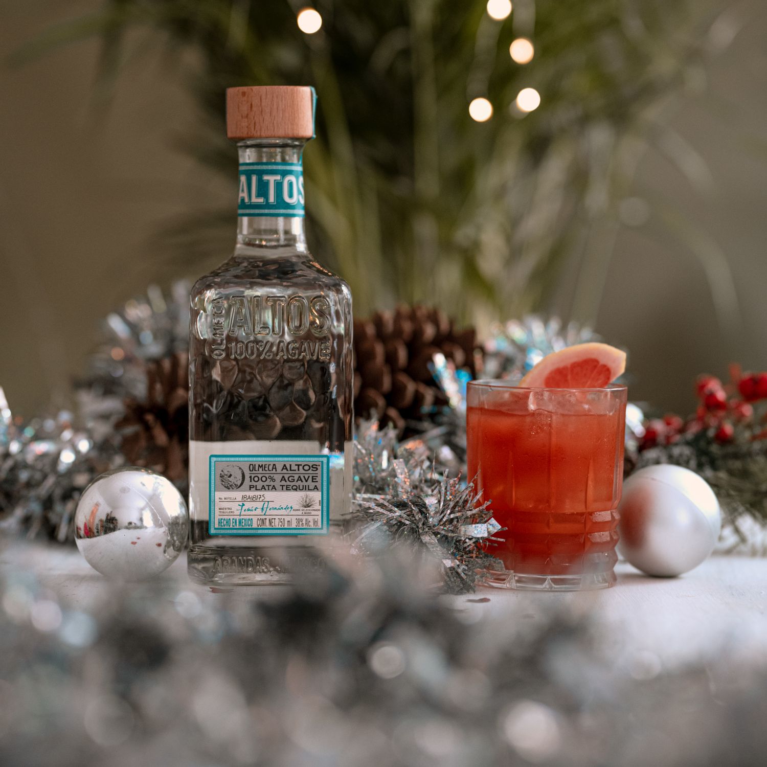 A bottle of Olmeca Altos tequila with a red cocktail drink arranged among silver Christmas ornaments and pine decorations.