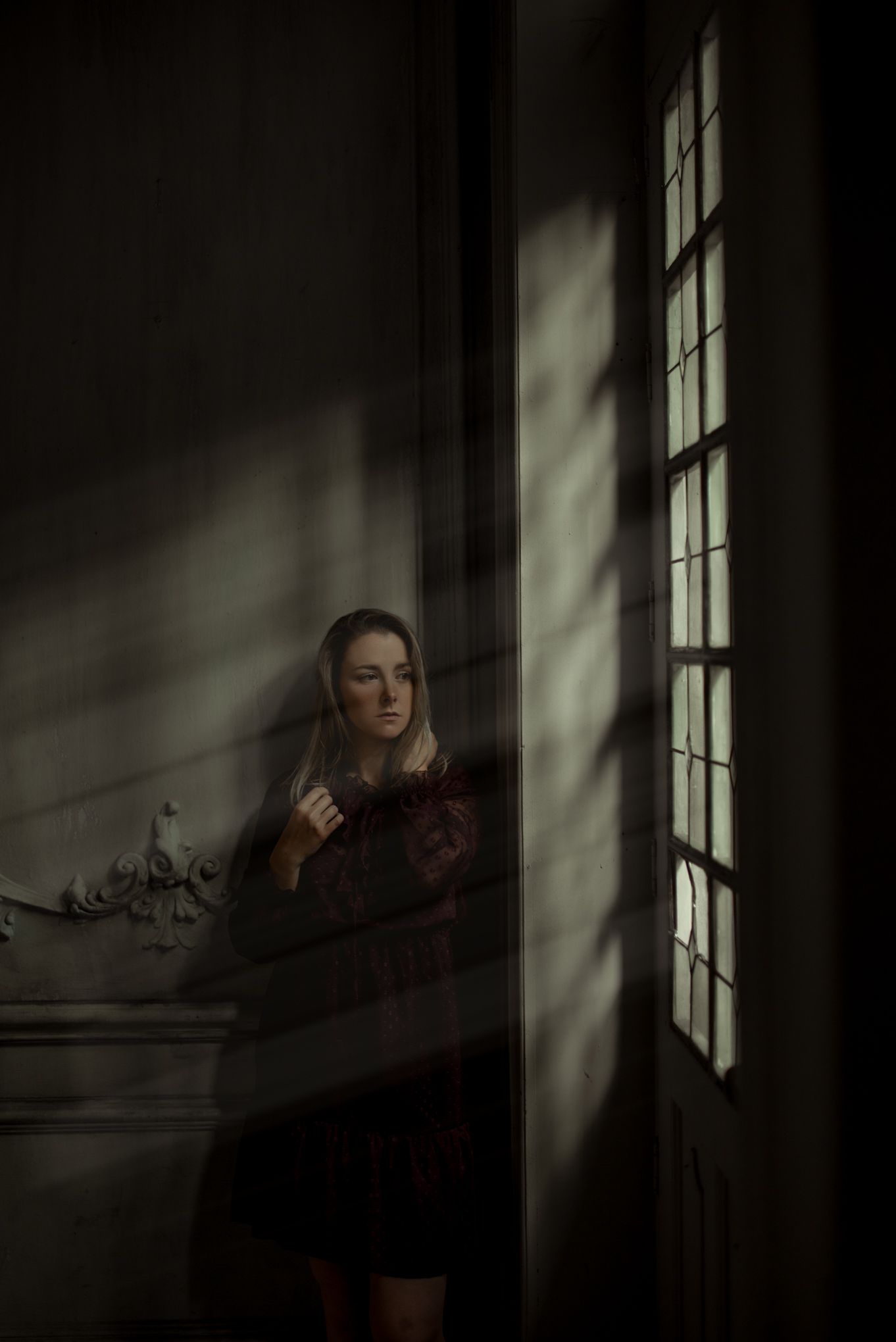 Artistic dark portrait by window with dramatic shadows and soft natural light flowing through panes.