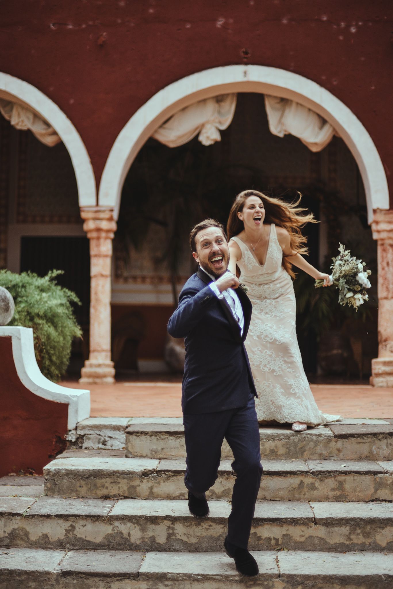 A joyful couple in wedding attire running down stone steps with white arches and a red wall in the background.