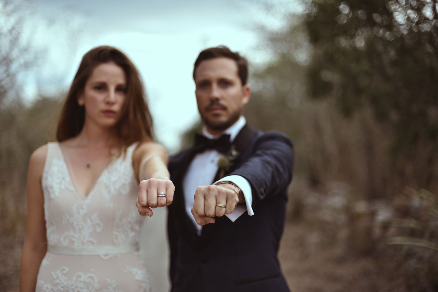 Two newlyweds show their wedding rings to the camera in a romantic outdoor setting with soft focus background.