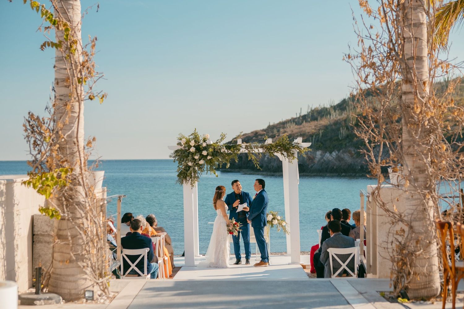 A scenic Mediterranean wedding ceremony takes place under a white arch overlooking the sea and cliffside on a sunny day.
