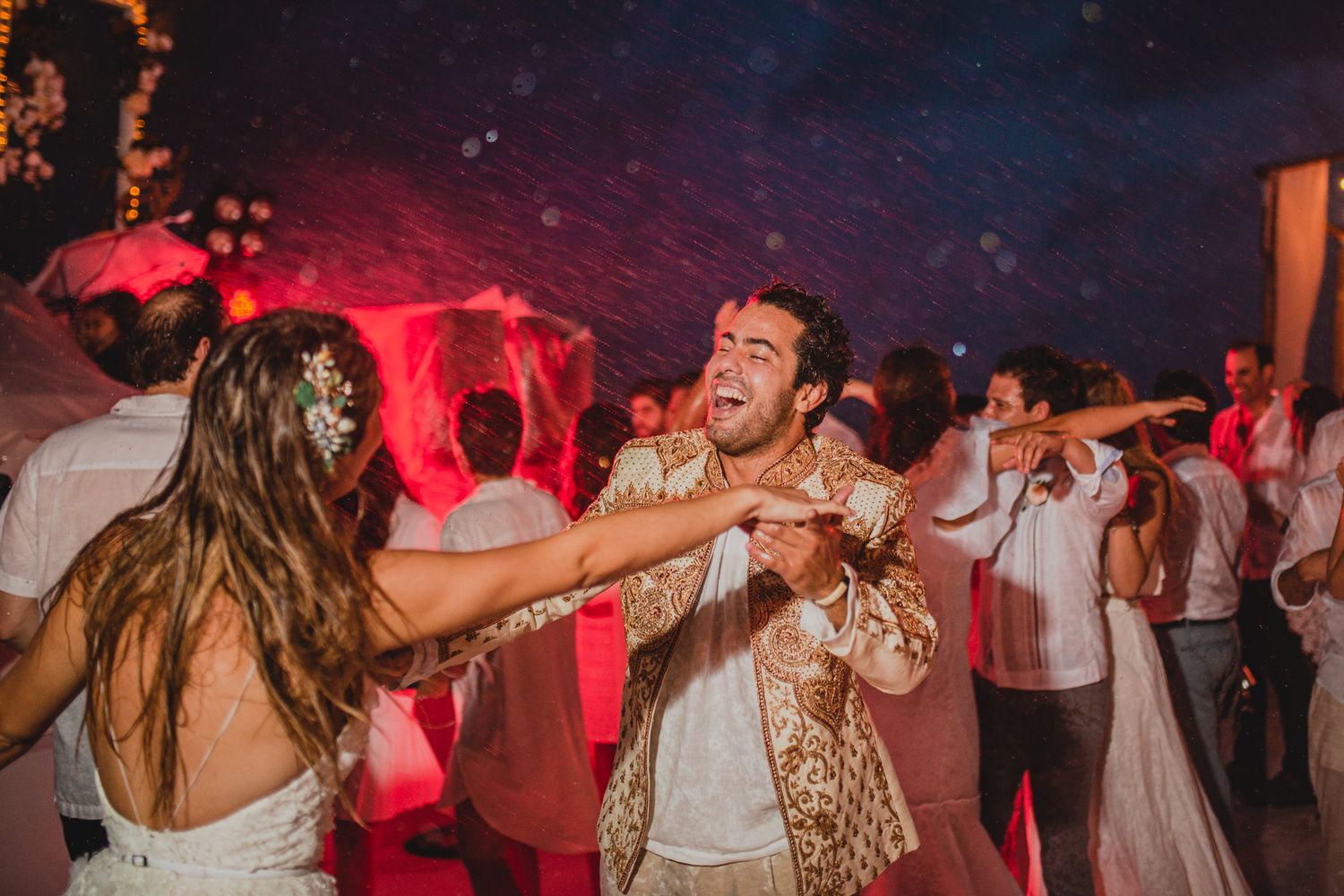 People dancing energetically at a nighttime wedding reception with red lighting and a festive atmosphere.