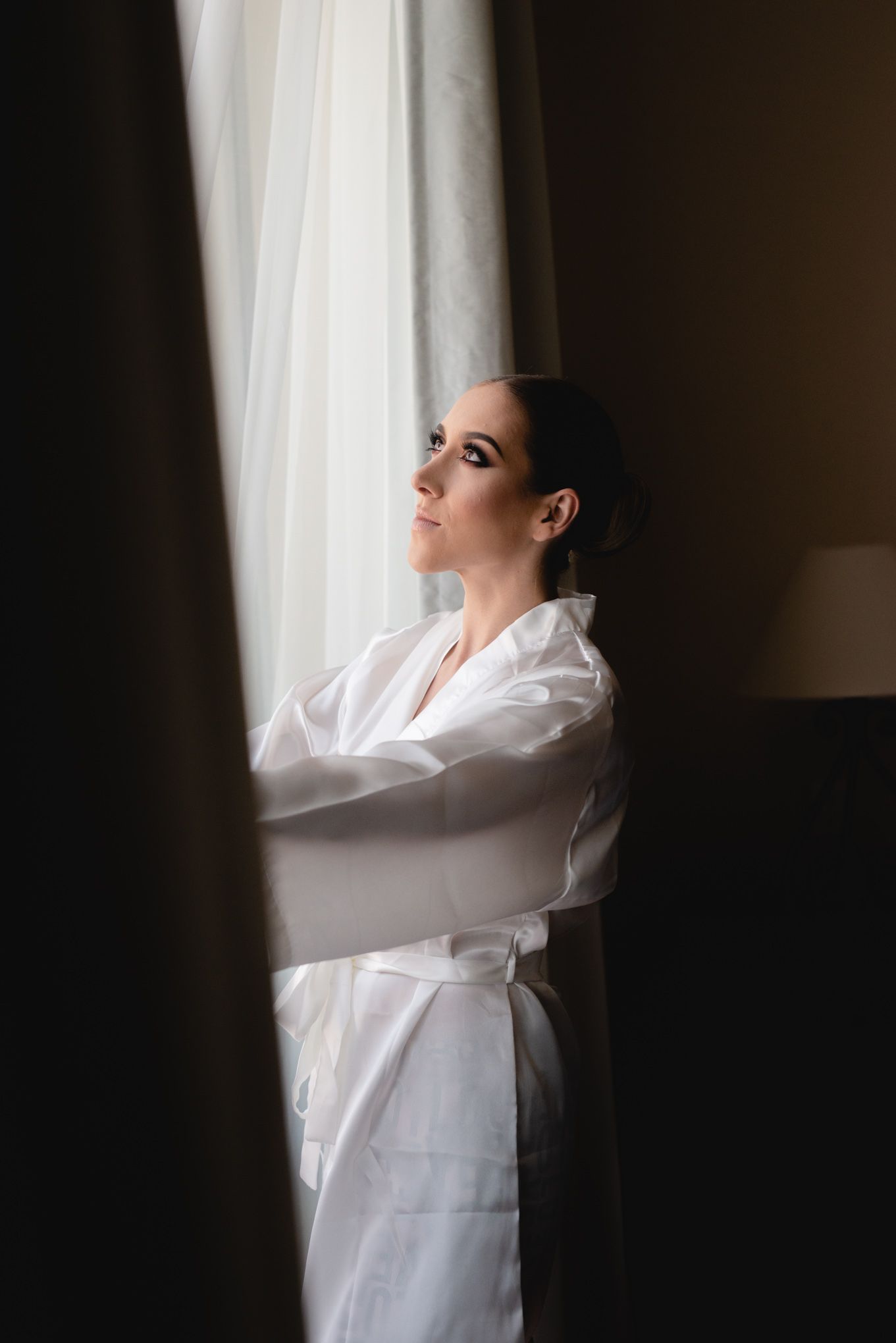 Someone in a white silk robe stands contemplatively by a window with soft natural lighting creating a moody atmosphere.