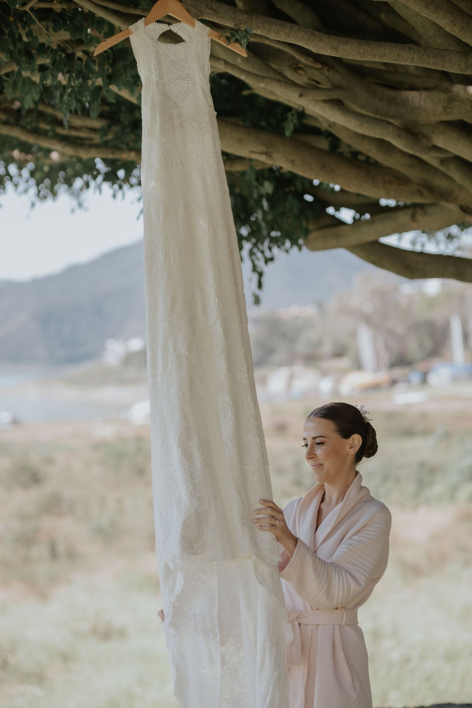 A bride in a robe admires her hanging wedding dress in natural morning light.