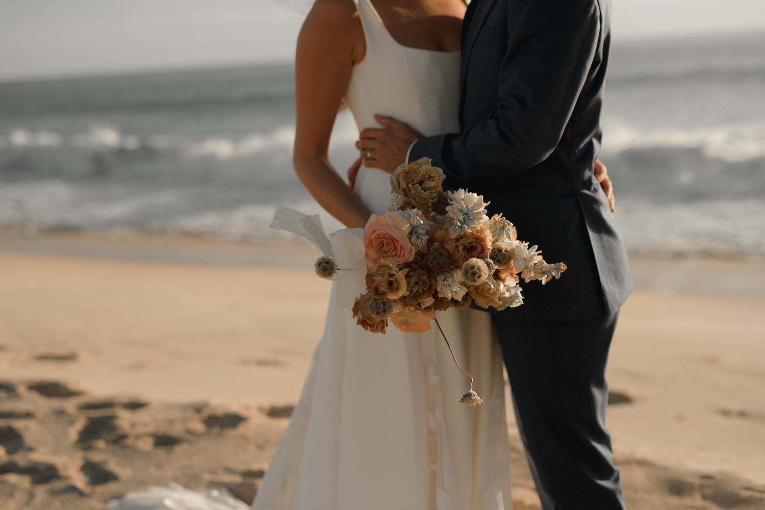 A couple holding dried flower bouquet stands together on sandy beach at sunset.