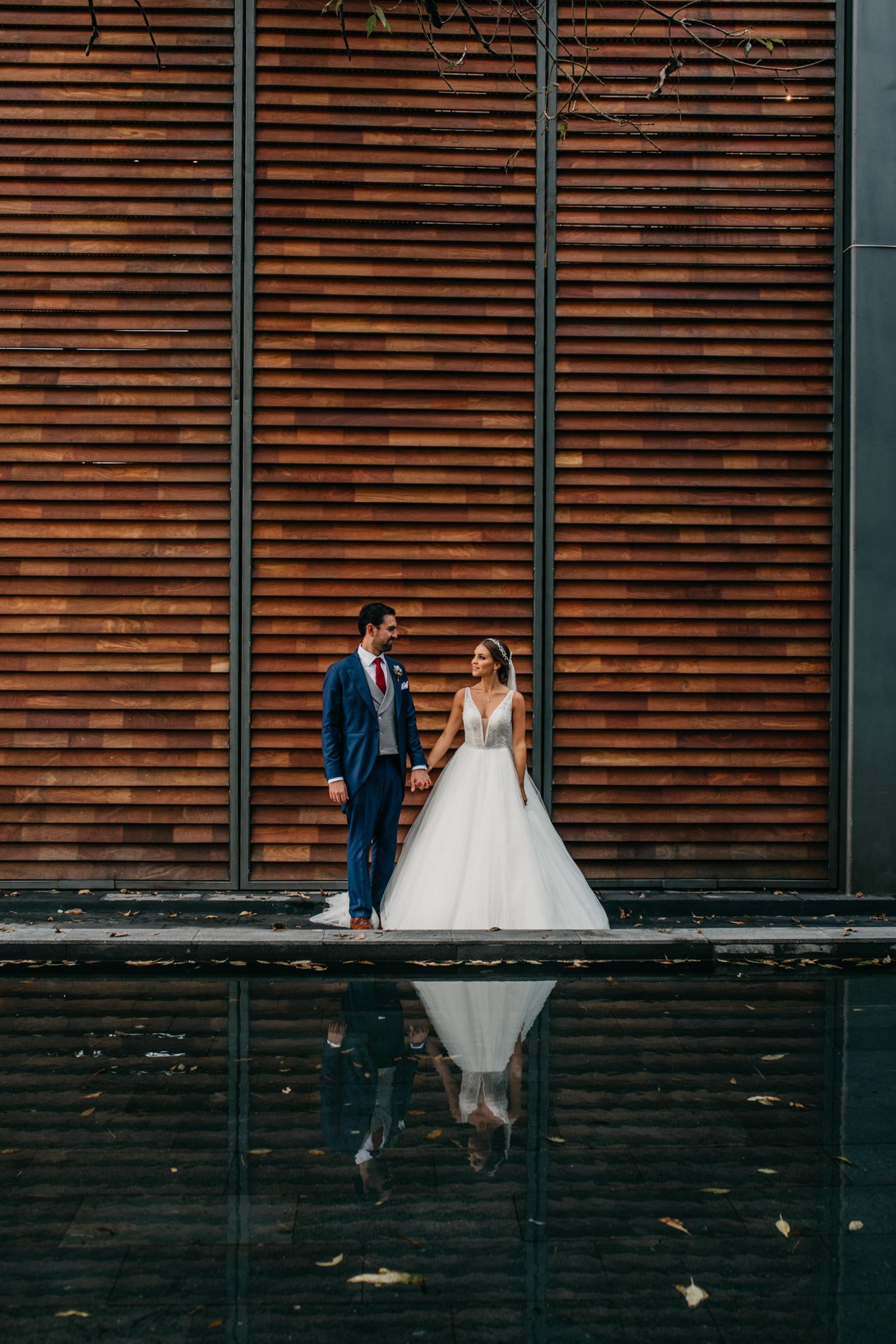 Wedding couple stands by wooden wall with perfect reflection in water below.