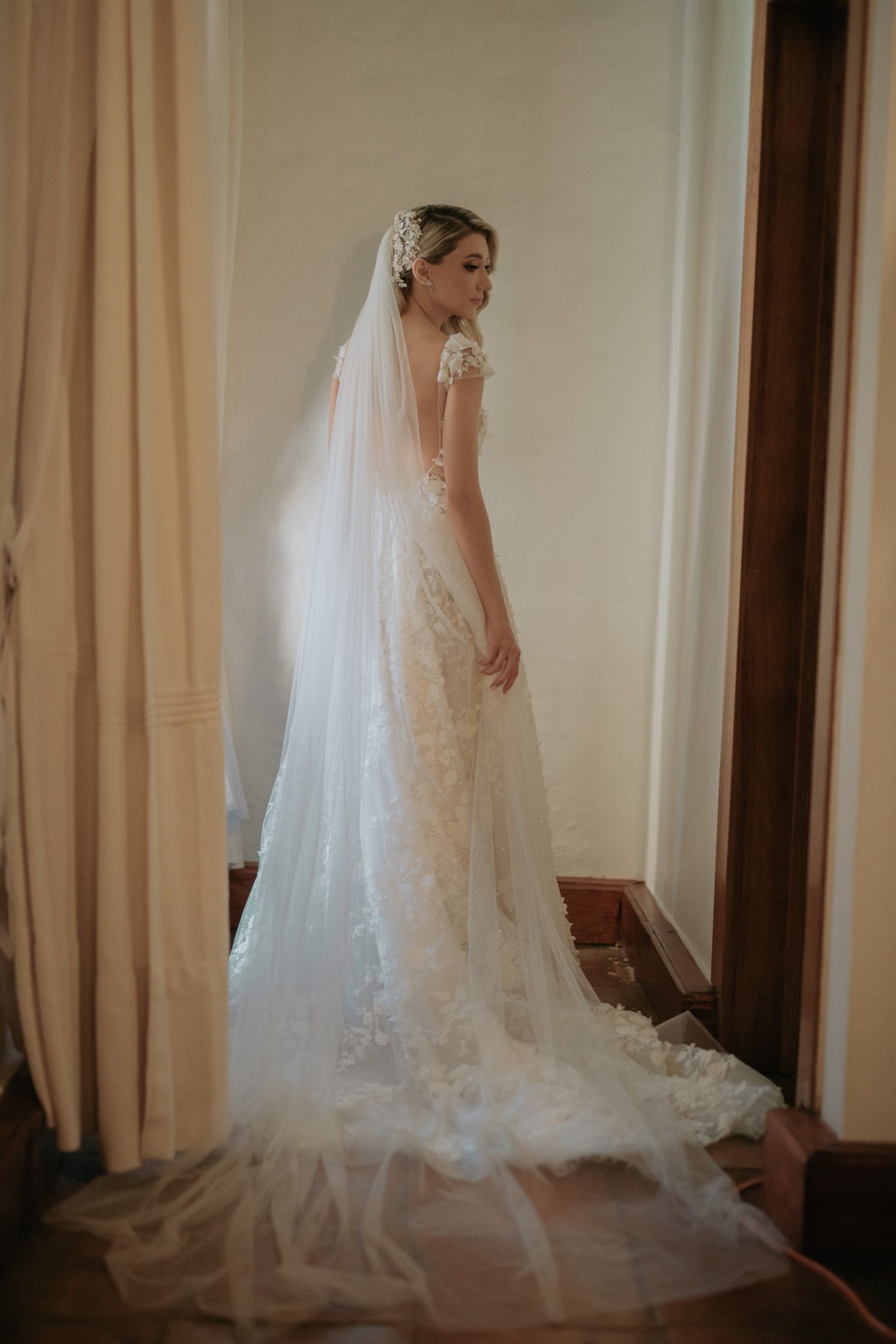 Bride in lace gown with flowing veil poses in elegant bridal portrait.