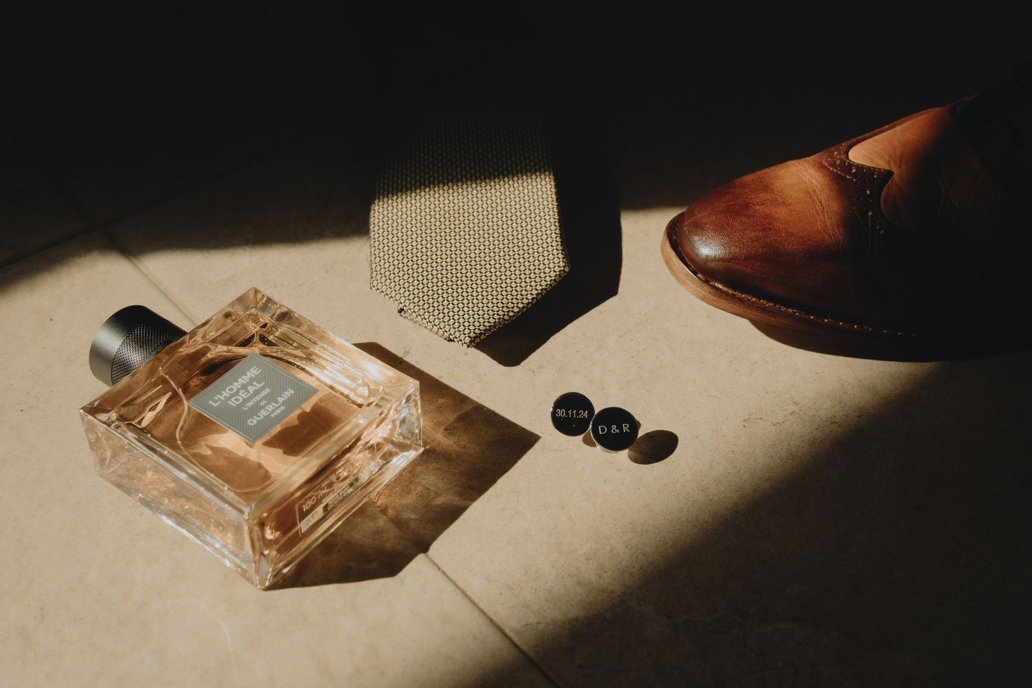 Perfume bottle and cufflinks arranged on beige surface in dramatic lighting.