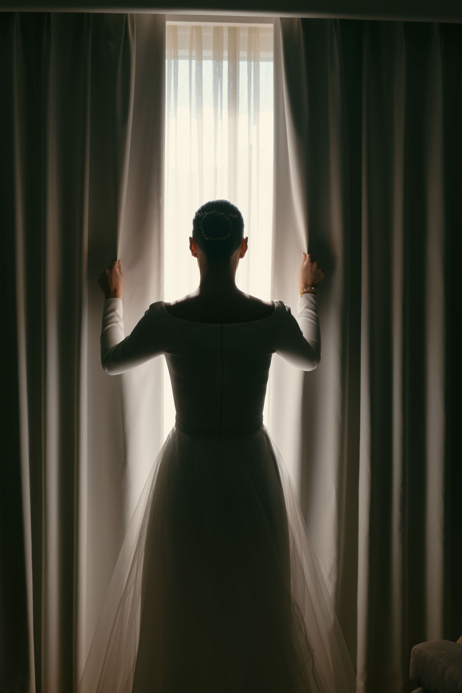 Silhouette of a person in formal dress opening curtains to reveal bright window light.