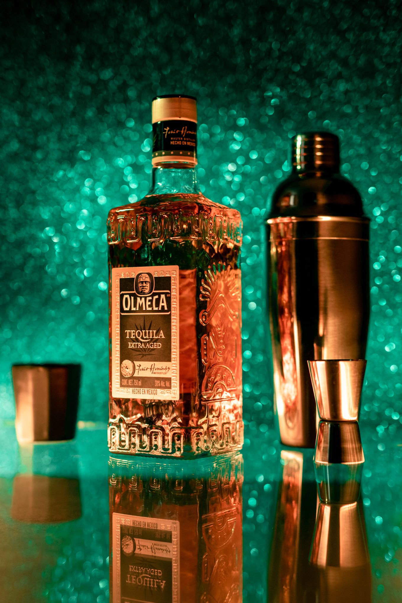 Olmeca tequila bottle and cocktail shaker displayed on reflective surface with green lighting.