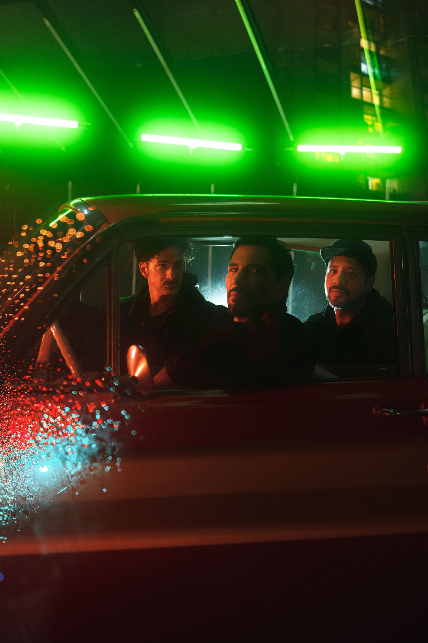 Three people sit in a car at night illuminated by bright green lights above while rain drops reflect on the window.