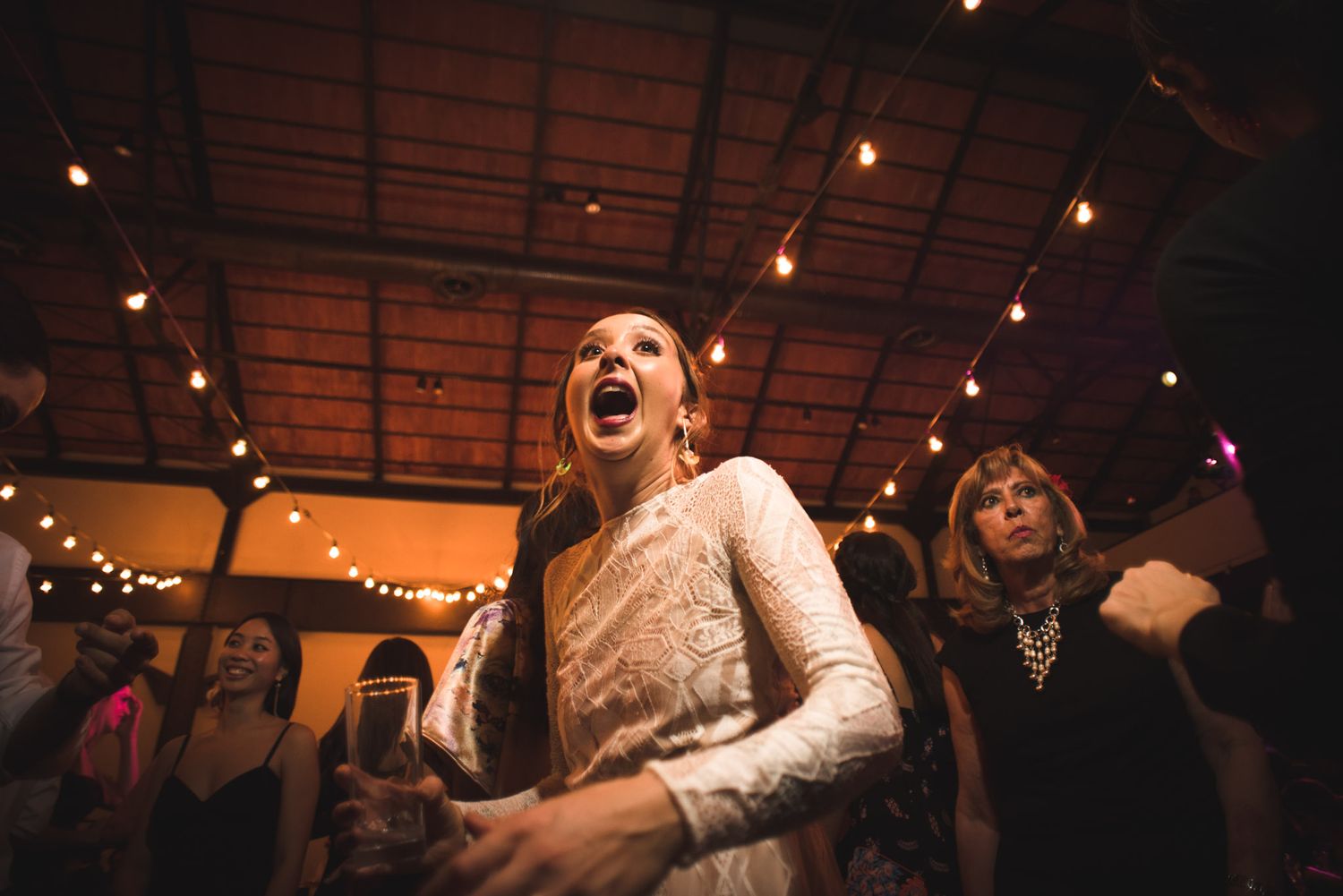 Glowing string lights illuminate a lively indoor reception space as someone in white lace celebrates with expressive joy.