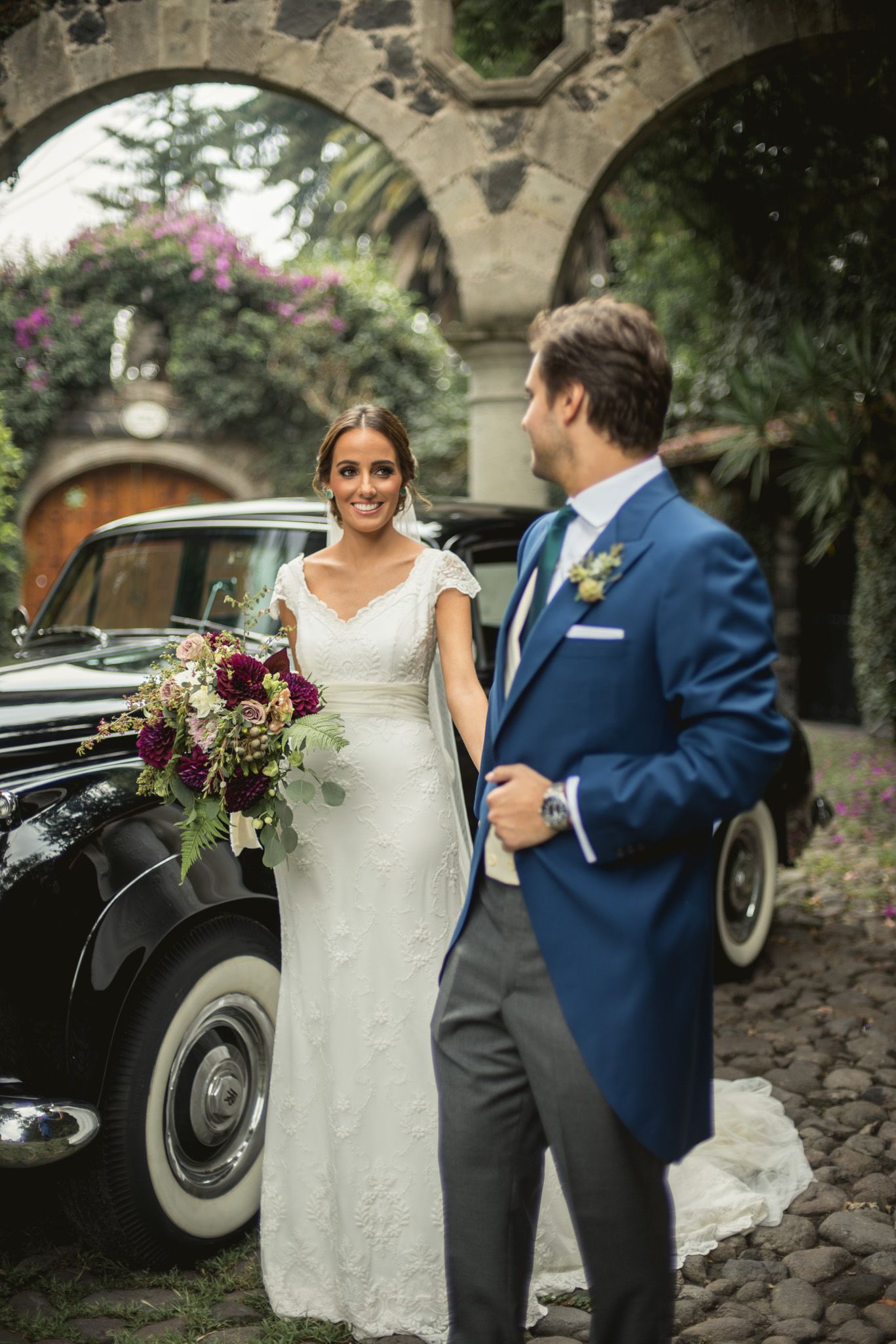 A newlywed couple poses by a vintage black car under a stone archway with purple flowers in the background.