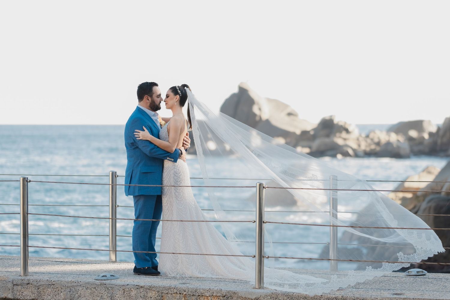 A couple in wedding attire embraces on an oceanfront terrace with waves crashing on rocky shore in background.