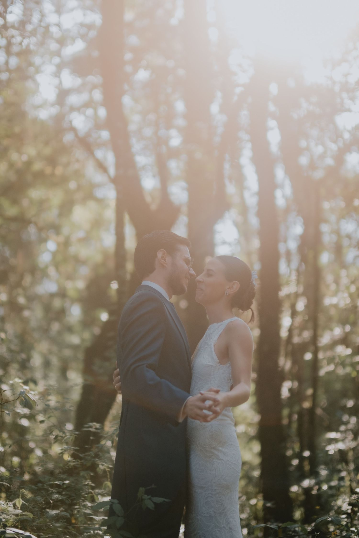 Newlyweds share intimate moment in sunlit forest setting.
