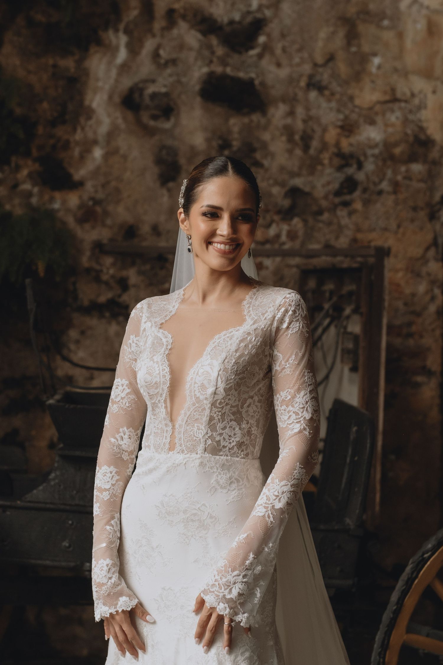 Radiant bride in long-sleeve lace wedding dress posing against textured wall.