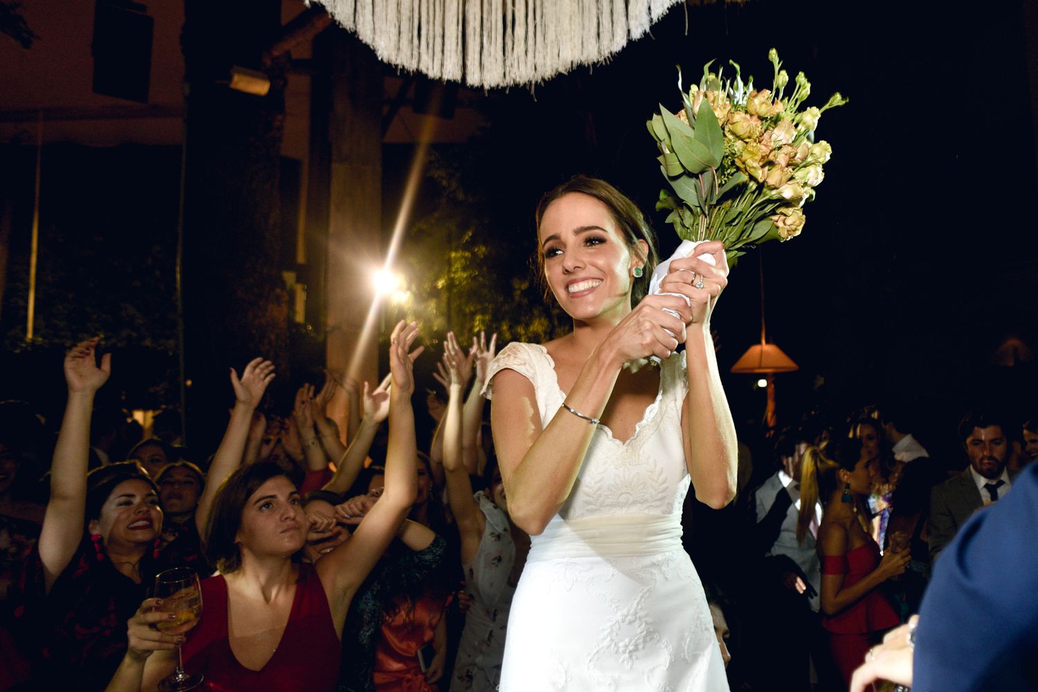 A bride holds up a bouquet while celebrating with wedding guests cheering behind her.