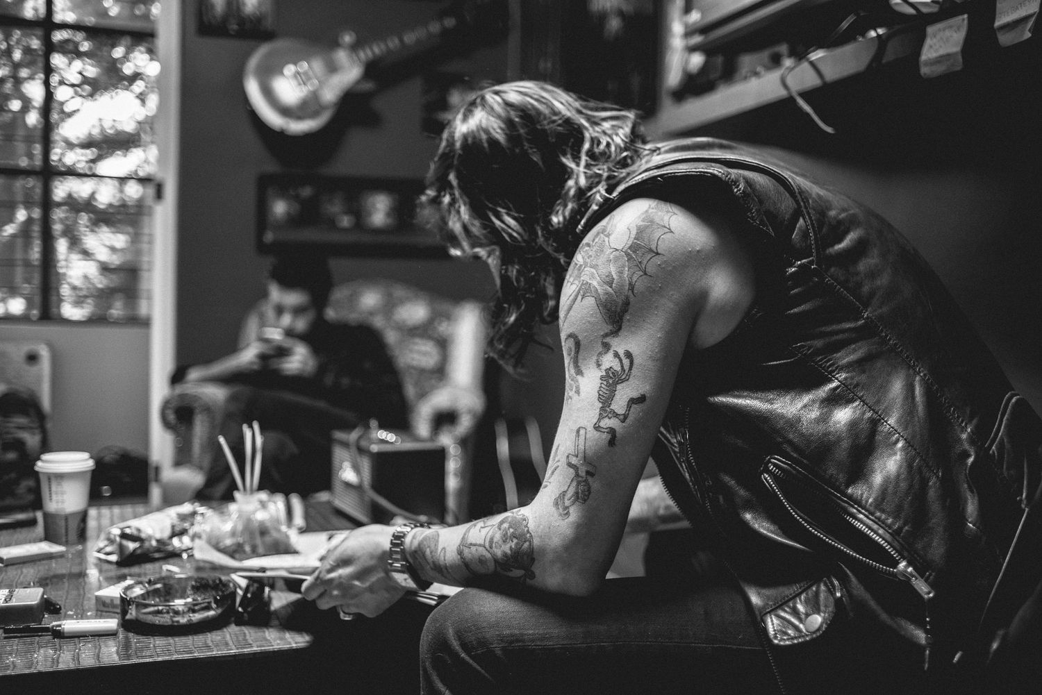 Tattooed musician sitting at table in dimly lit bar with guitars visible on wall.