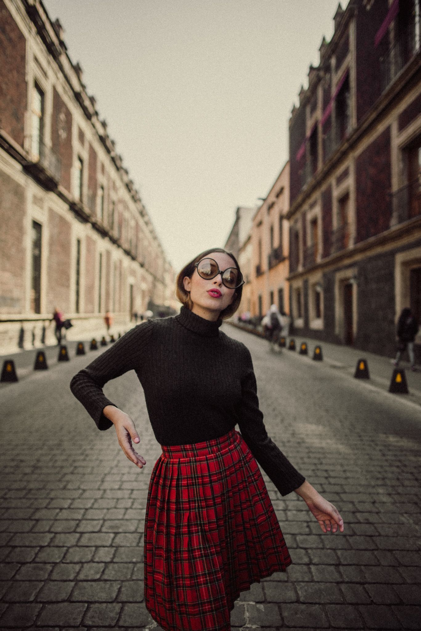 Retro street style with black turtleneck and red plaid skirt on cobblestone street.