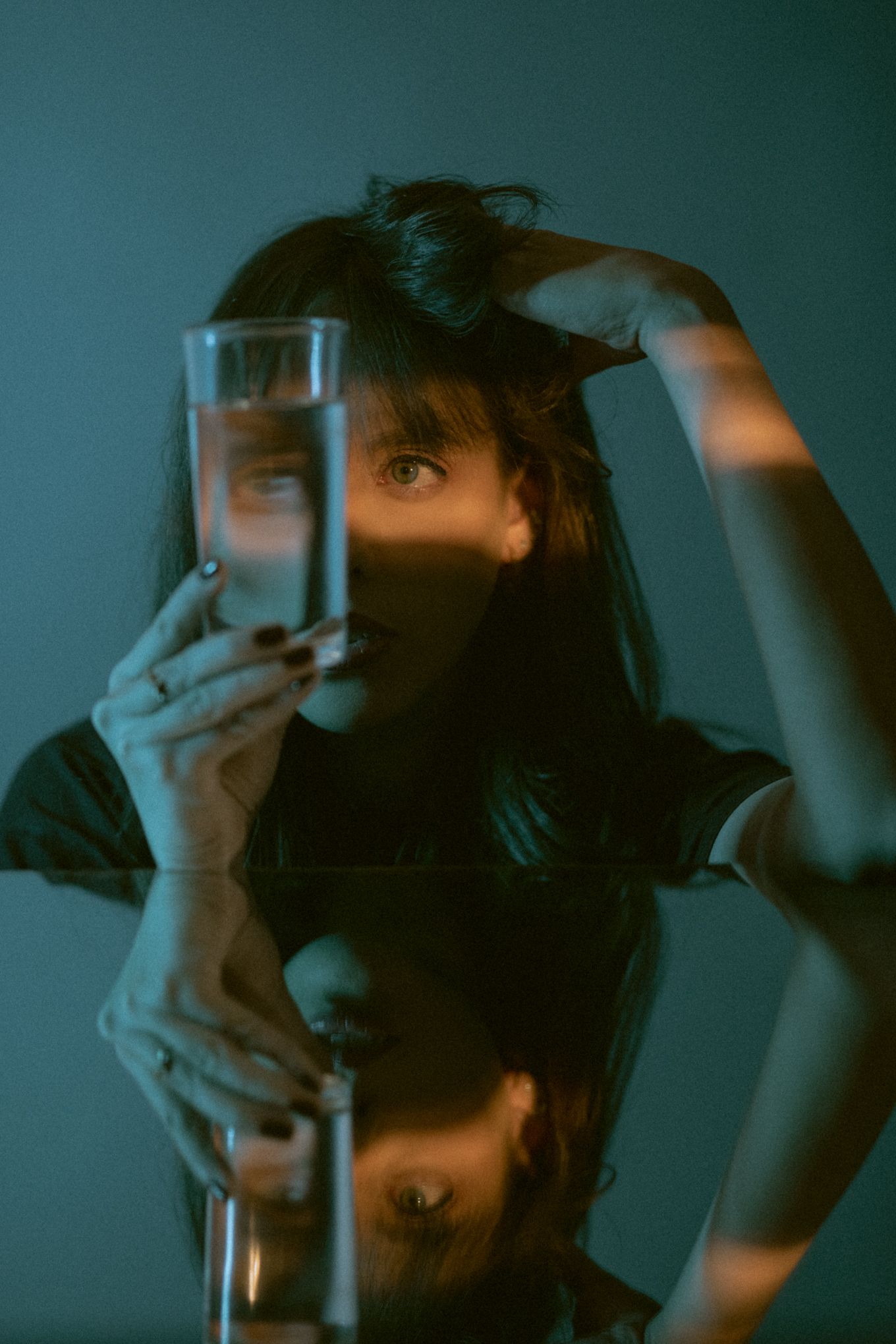 Moody portrait with a glass of water in dark teal lighting creating dramatic reflections and shadows.