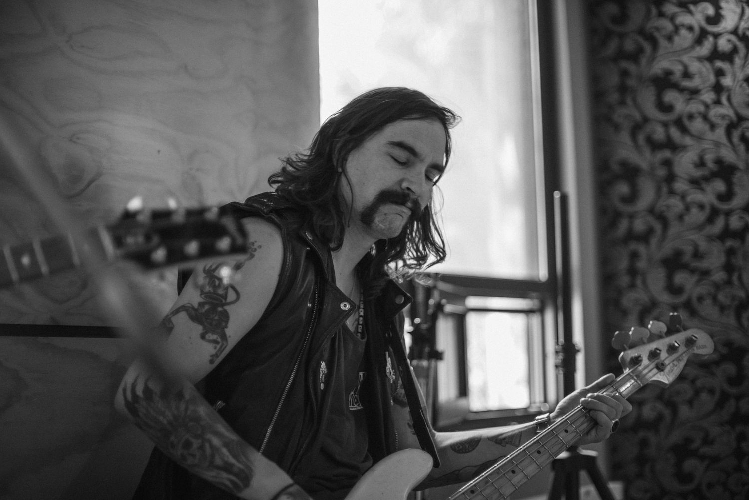 Musician plays bass guitar in black and white photograph with moody lighting and ornate wall patterns.