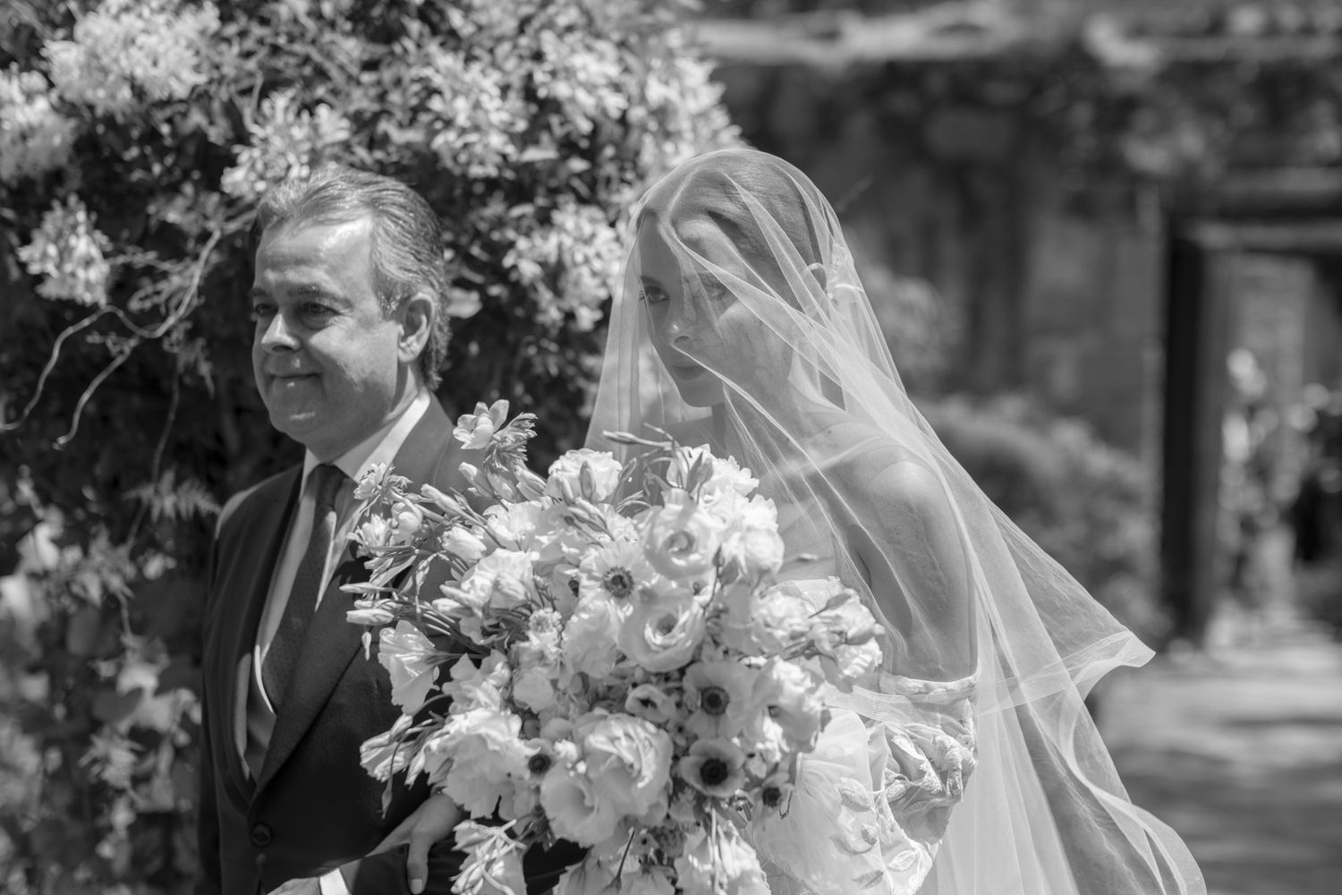 Black and white wedding photo capturing a walk down the aisle with a lush floral bouquet.