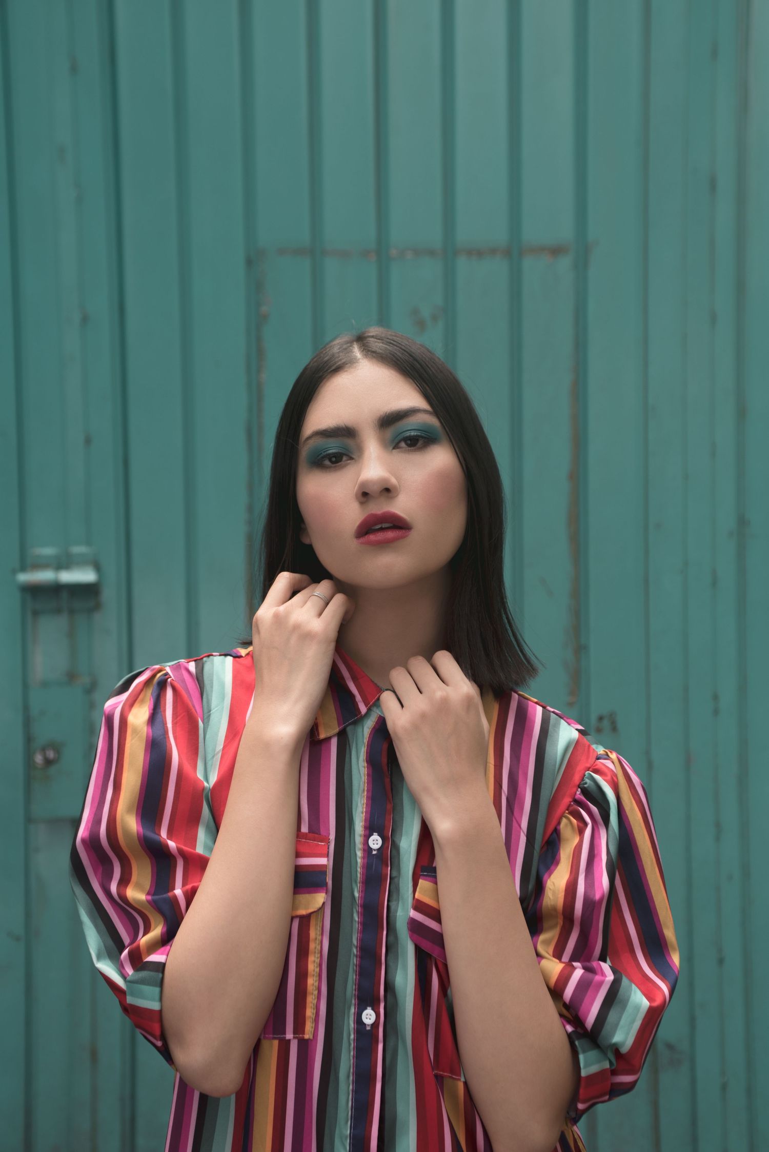 Fashion portrait with bold makeup against turquoise metal wall wearing striped shirt.