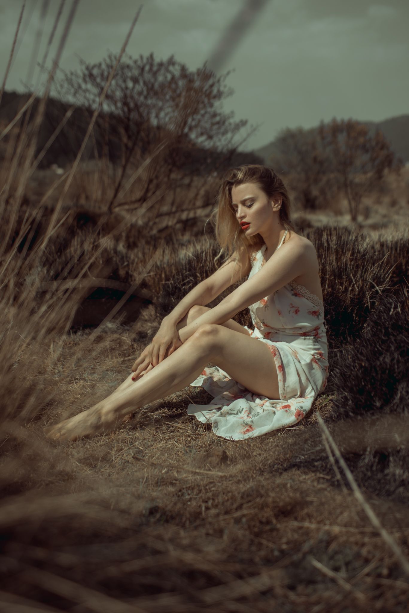 Woman in floral dress sits contemplatively on dry grass in moody vintage-style photo.