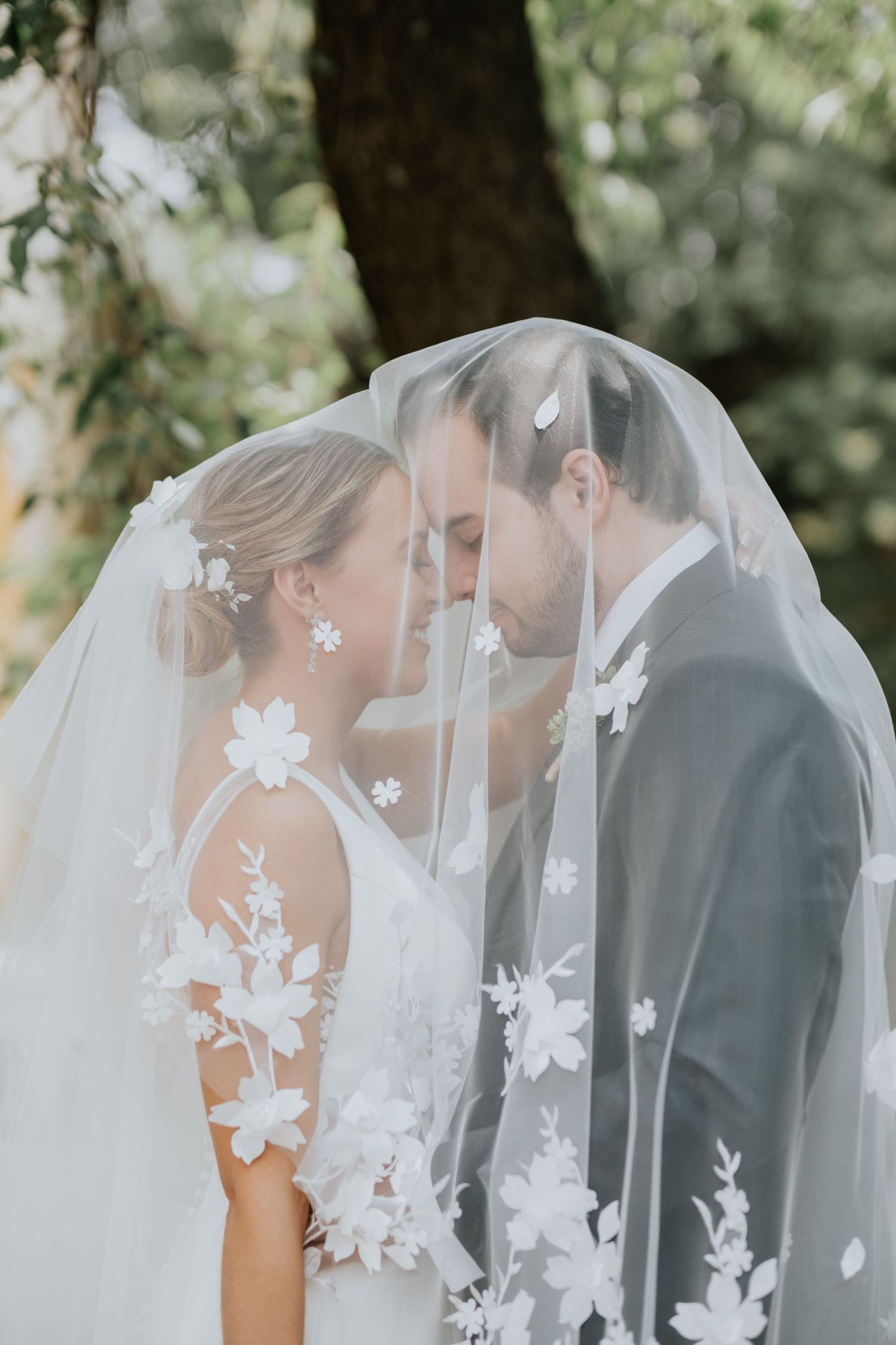 Romantic wedding moment captured under delicate floral veil with white lace appliques in outdoor garden setting.