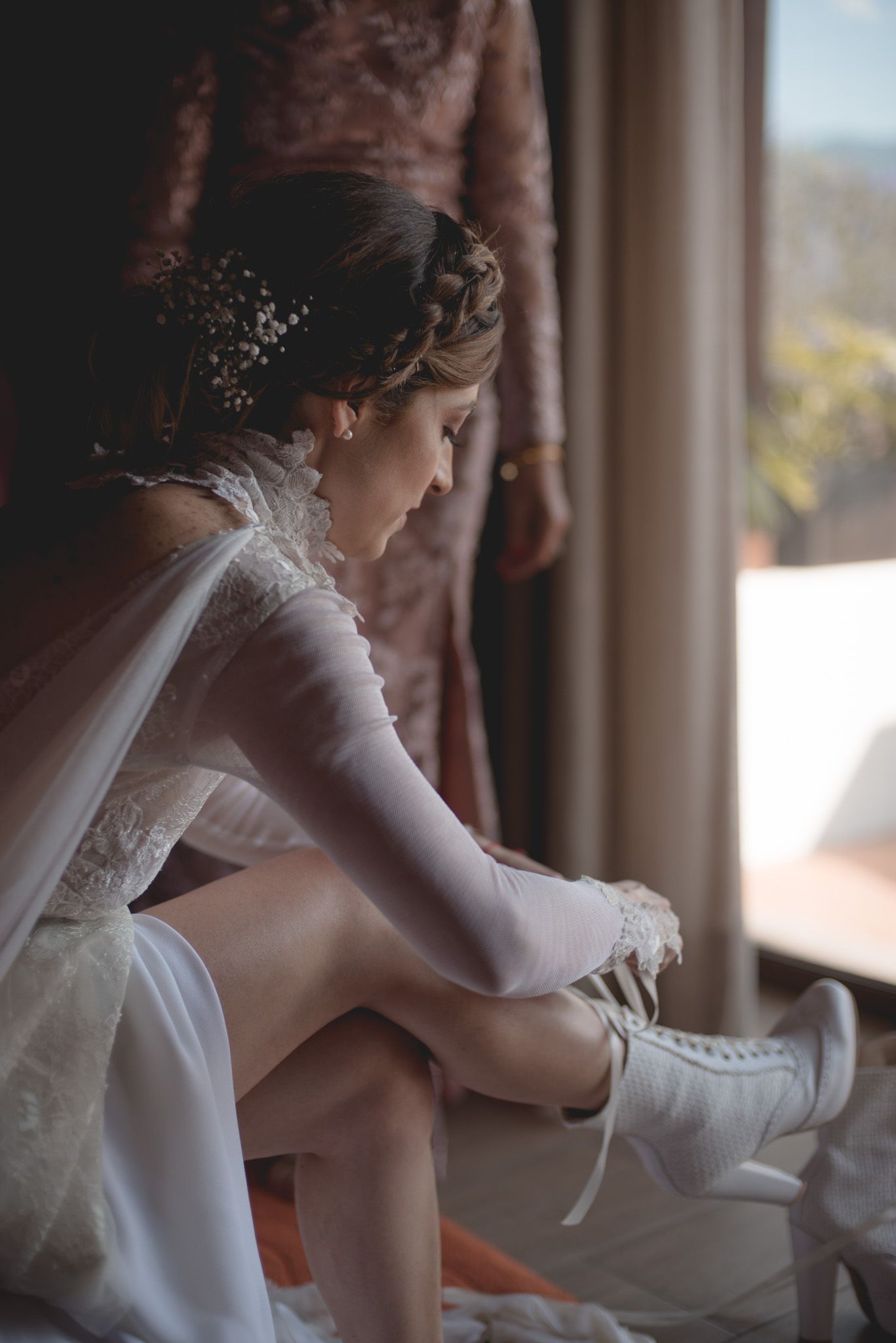 A bride in a white wedding dress and veil puts on elegant white shoes while getting ready by a window.