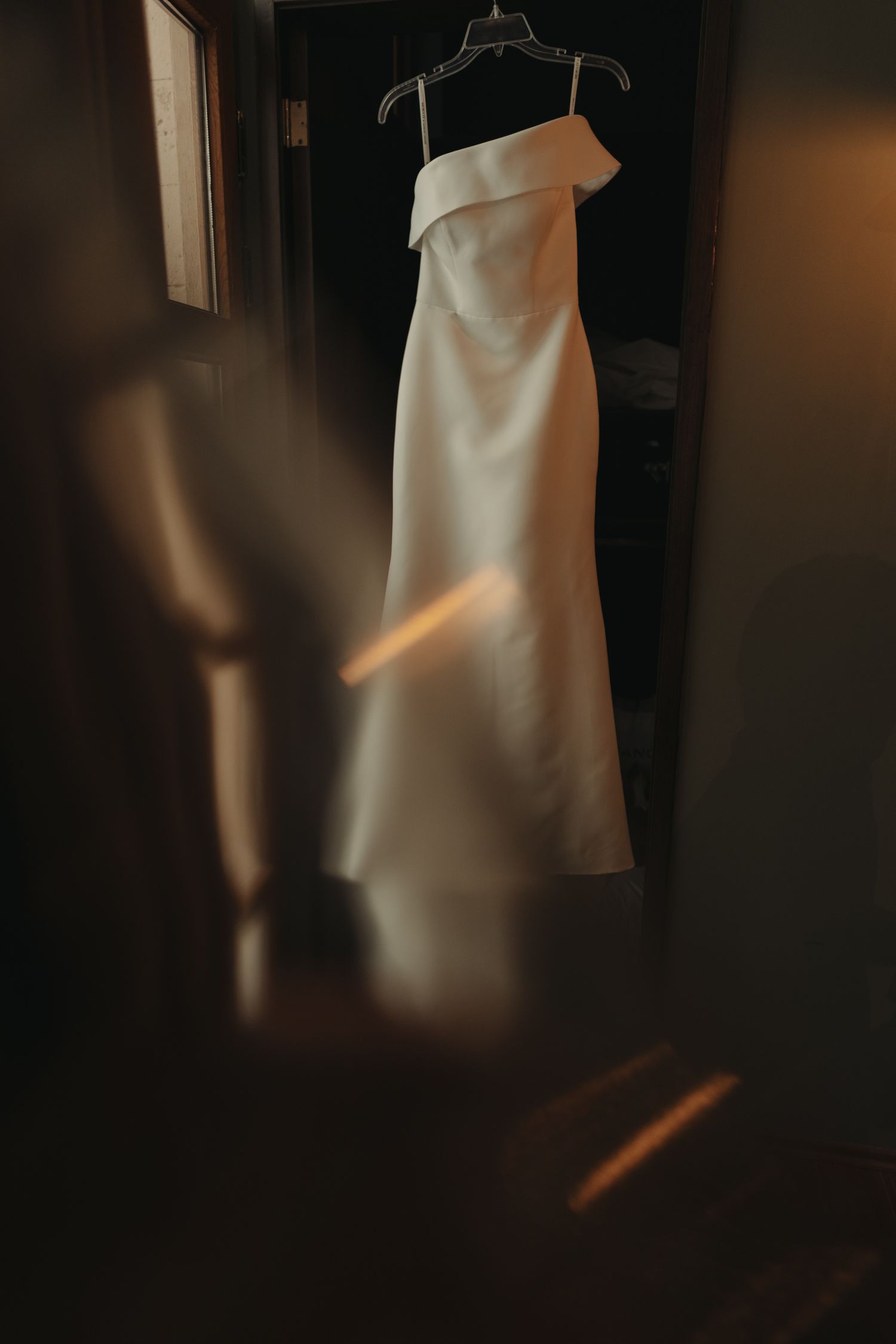 An elegant white one-shoulder wedding dress hangs on a black hanger against a dark moody backdrop with dramatic shadows.
