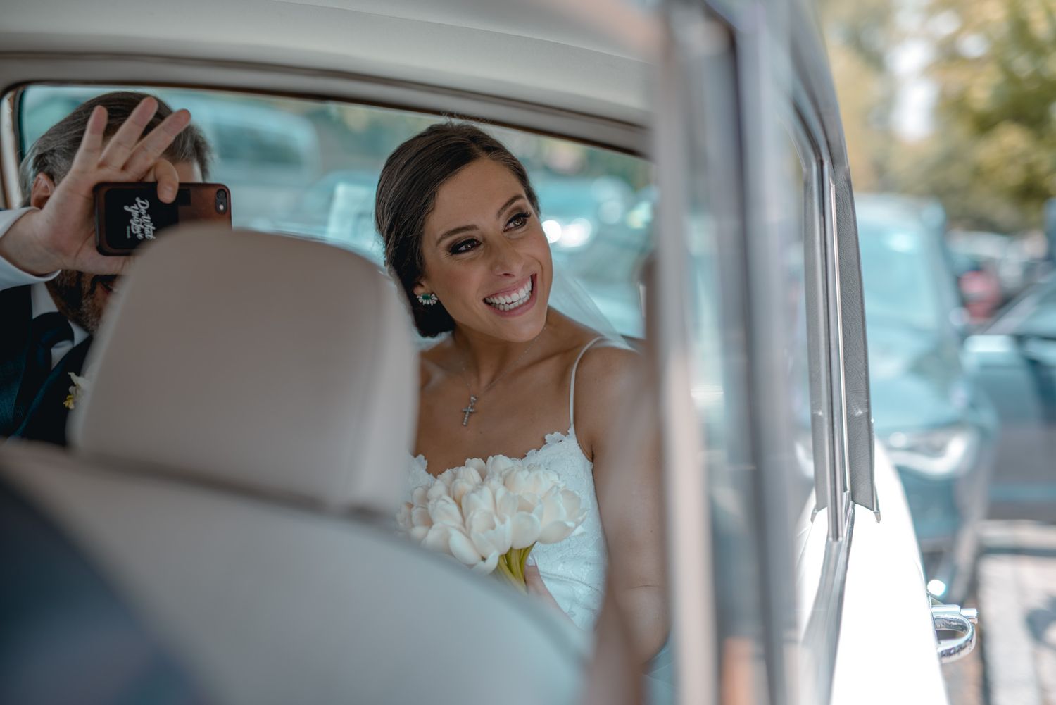 A smiling bride in a white dress sits in a car with white leather seats on her wedding day.