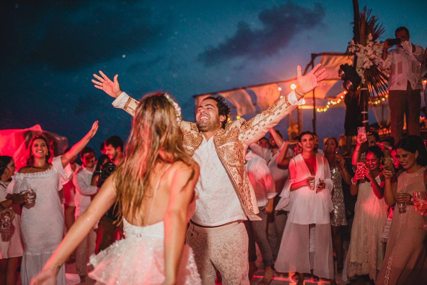 People dancing and celebrating at a evening beach party with red lighting and a festive atmosphere.