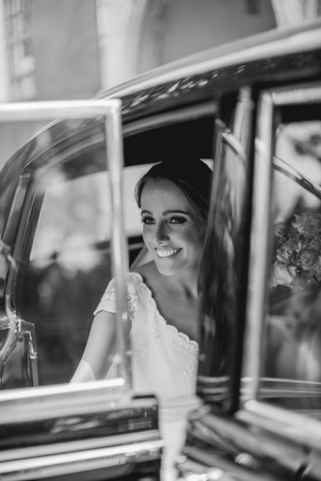 Bride in white lace dress smiles from vintage car window in black and white wedding photo.