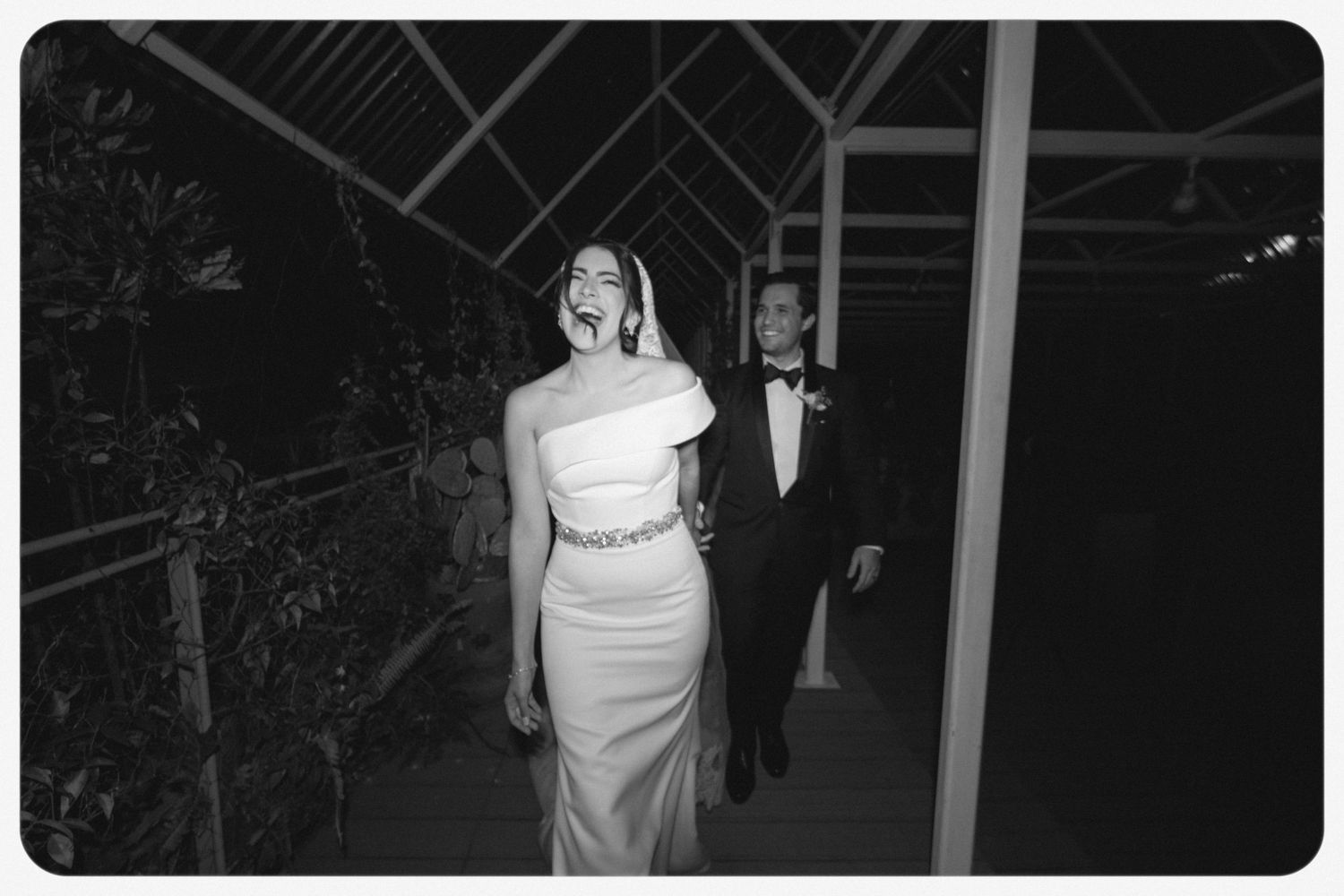 Black and white image of a formal event with a couple walking, woman wearing an elegant one-shoulder white gown.