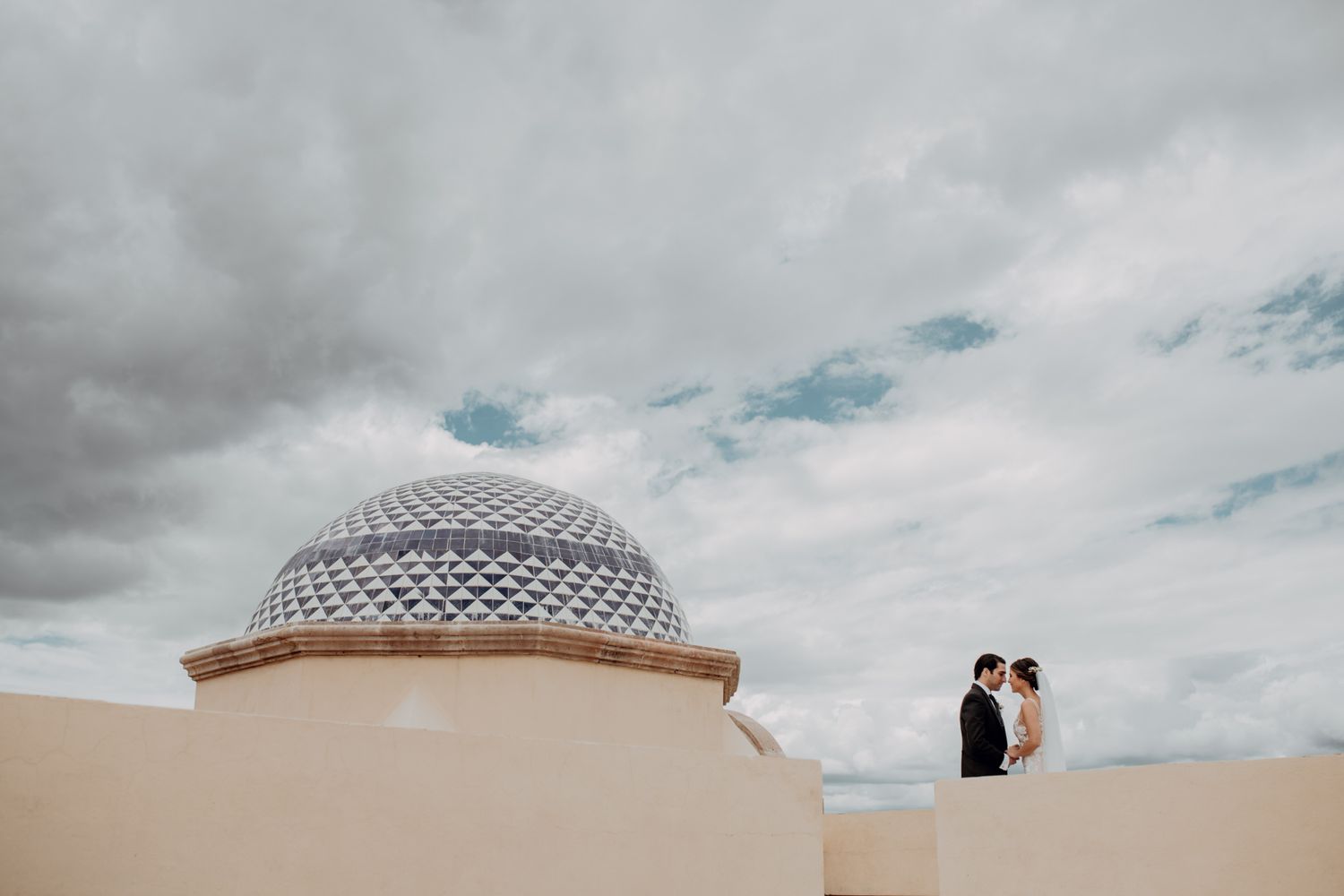 Two silhouettes stand atop a beige building with a domed structure against a cloudy sky.