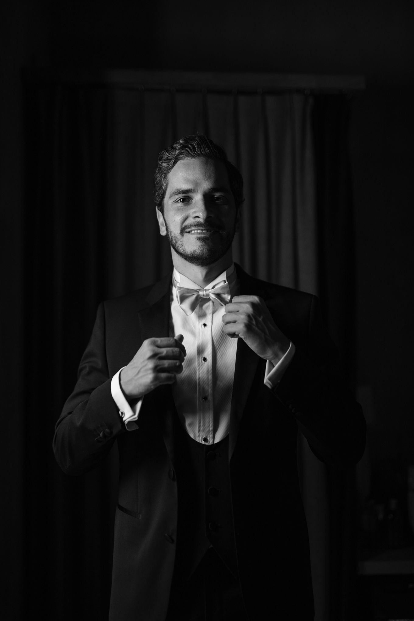 Groom adjusts bow tie while wearing classic black tuxedo in dramatic black and white portrait.