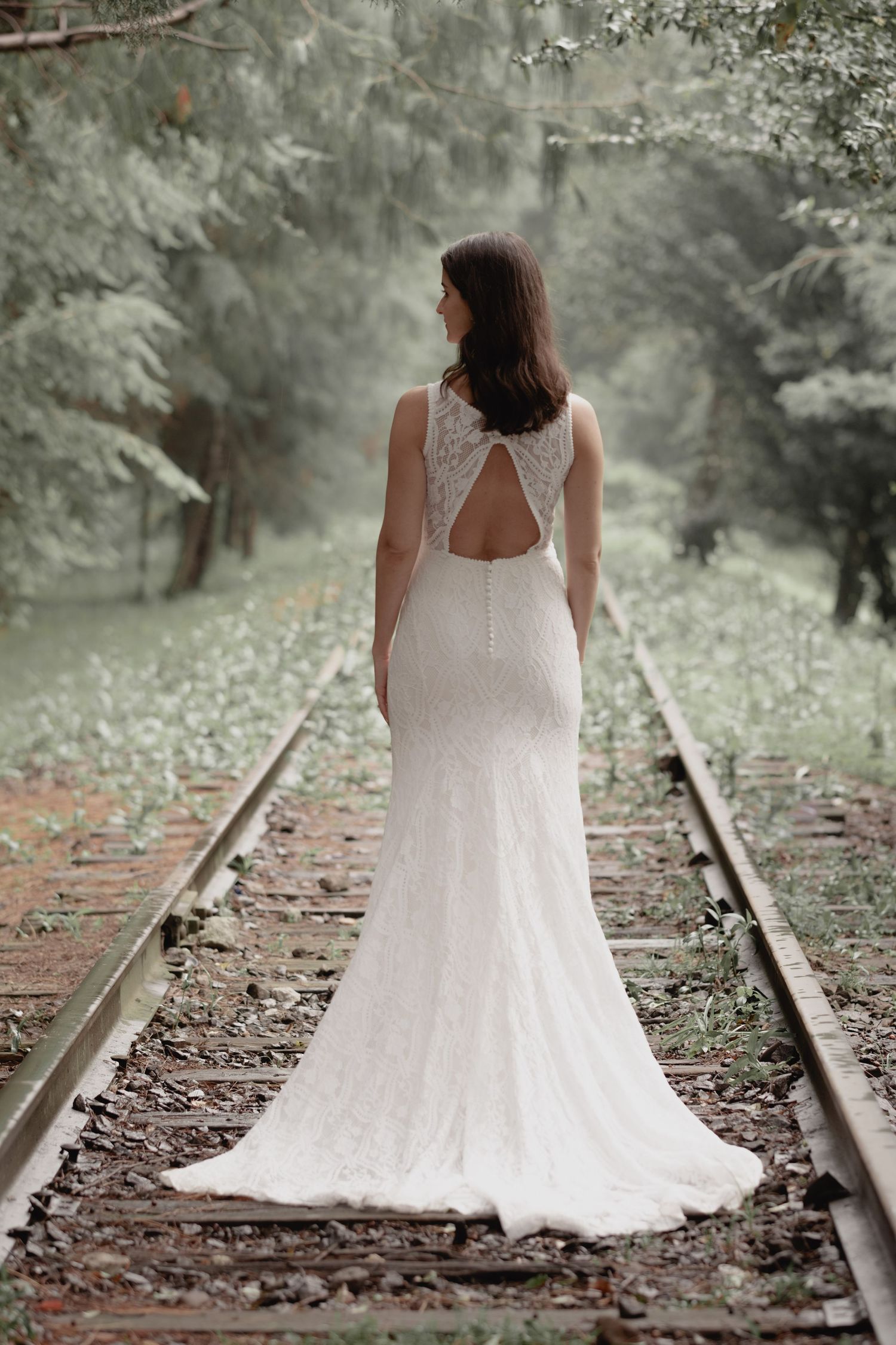A bride in a fitted lace wedding gown with keyhole back detail stands on train tracks in a misty forest setting.