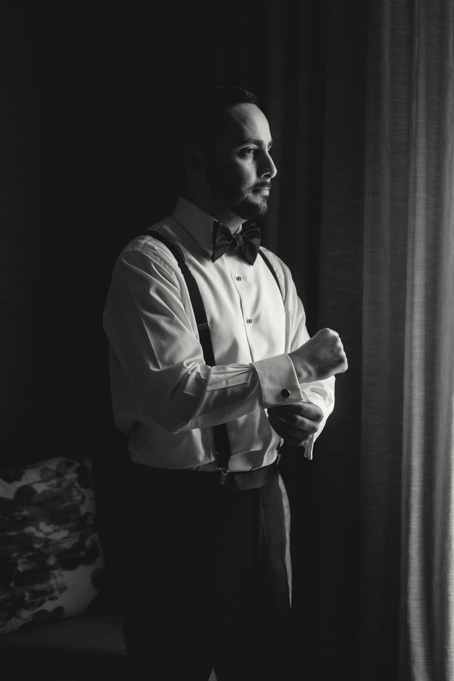 Moody black and white portrait of person adjusting cufflinks by window.