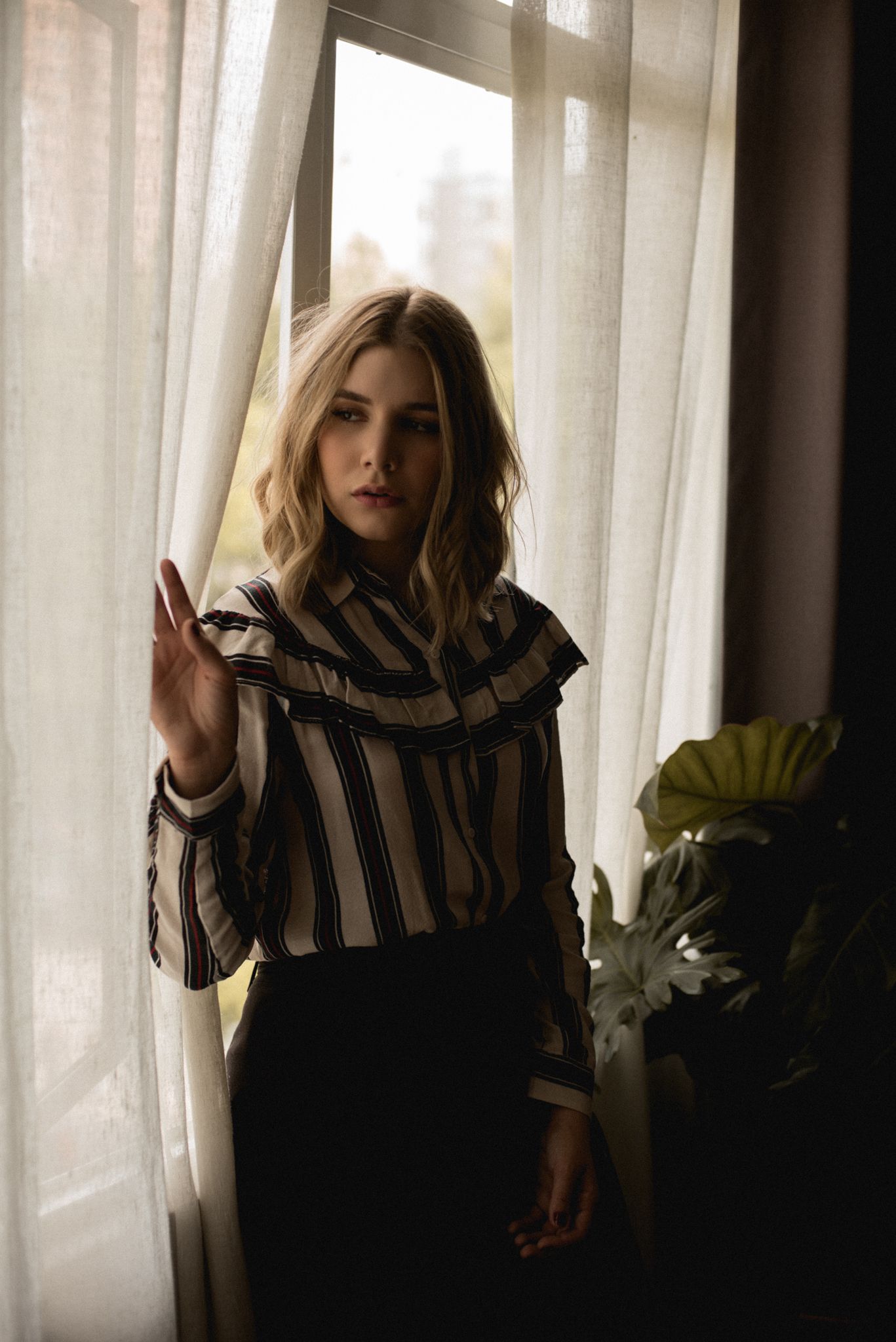 Moody portrait of woman in vintage striped top against sheer curtains.