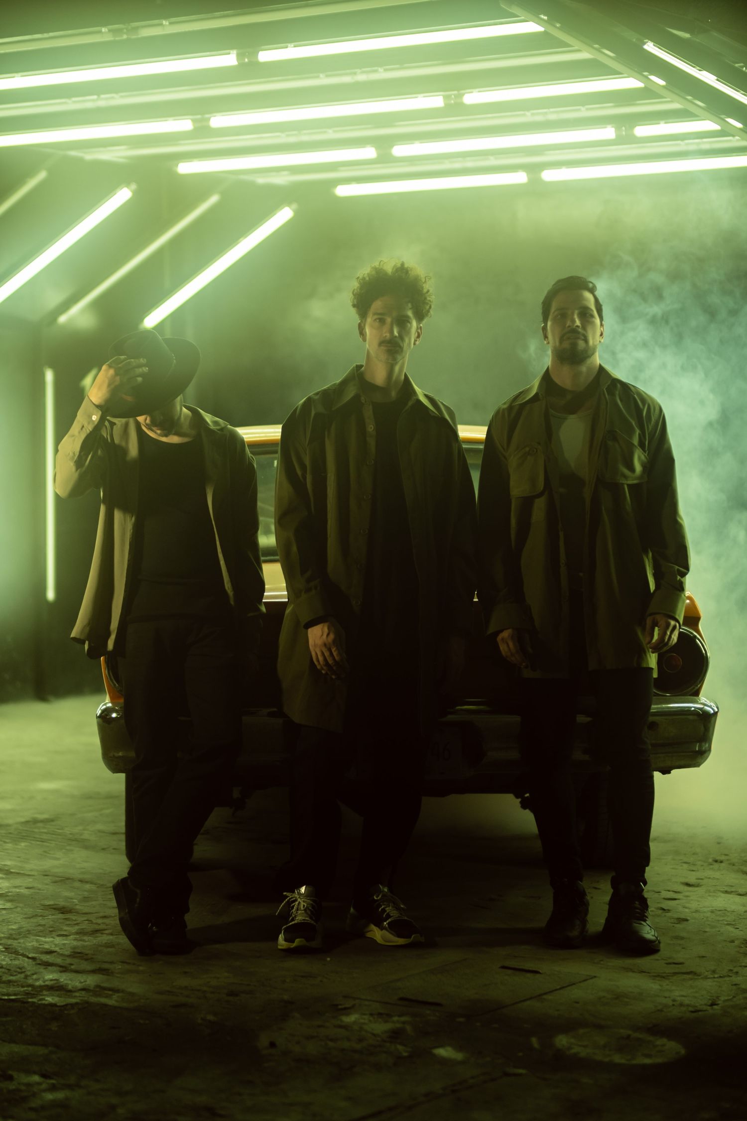 Three silhouetted figures stand in front of a car under dramatic green fluorescent lighting in a smoky atmosphere.
