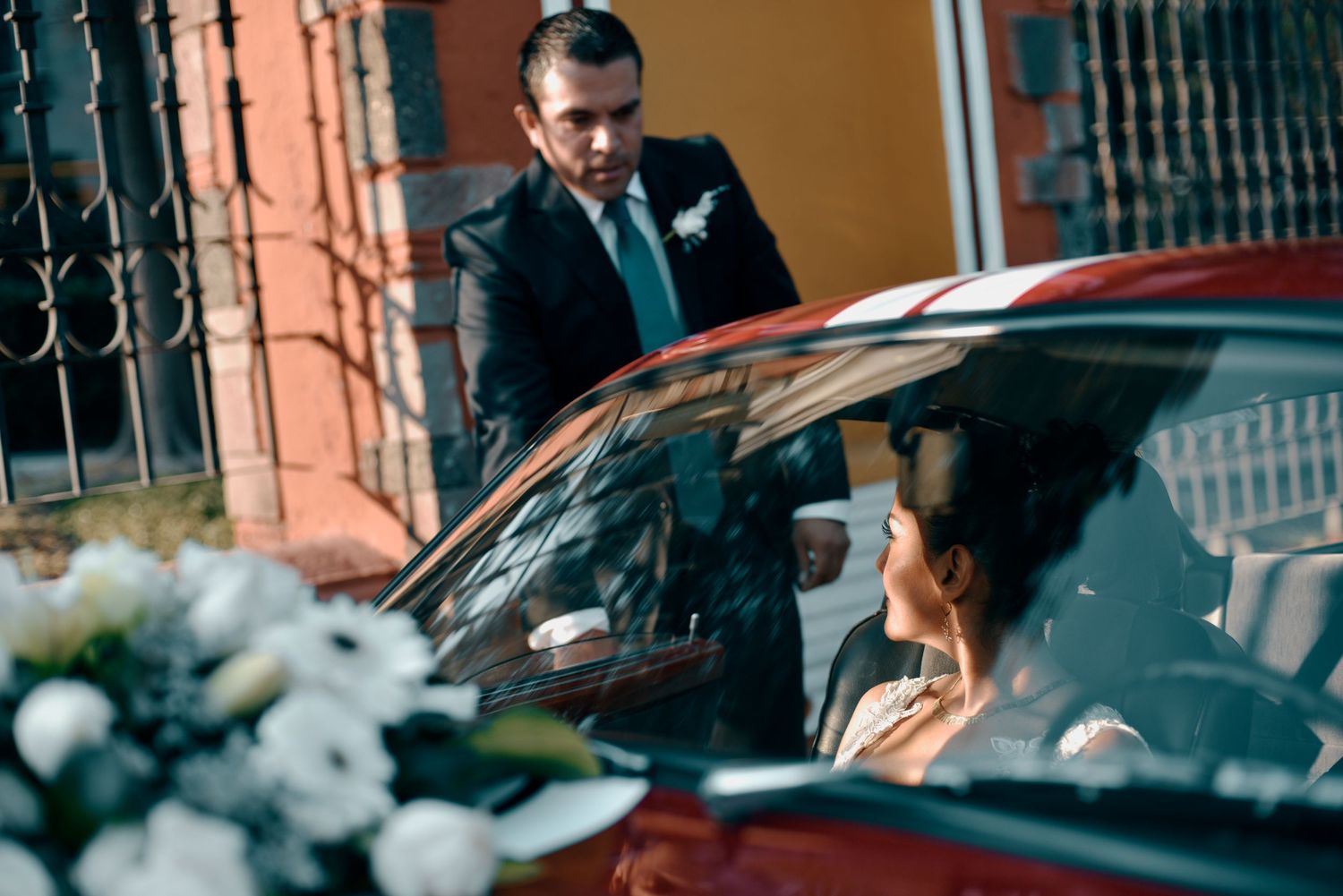 A well-dressed driver in a suit opens a car door for someone seated in a red luxury vehicle.