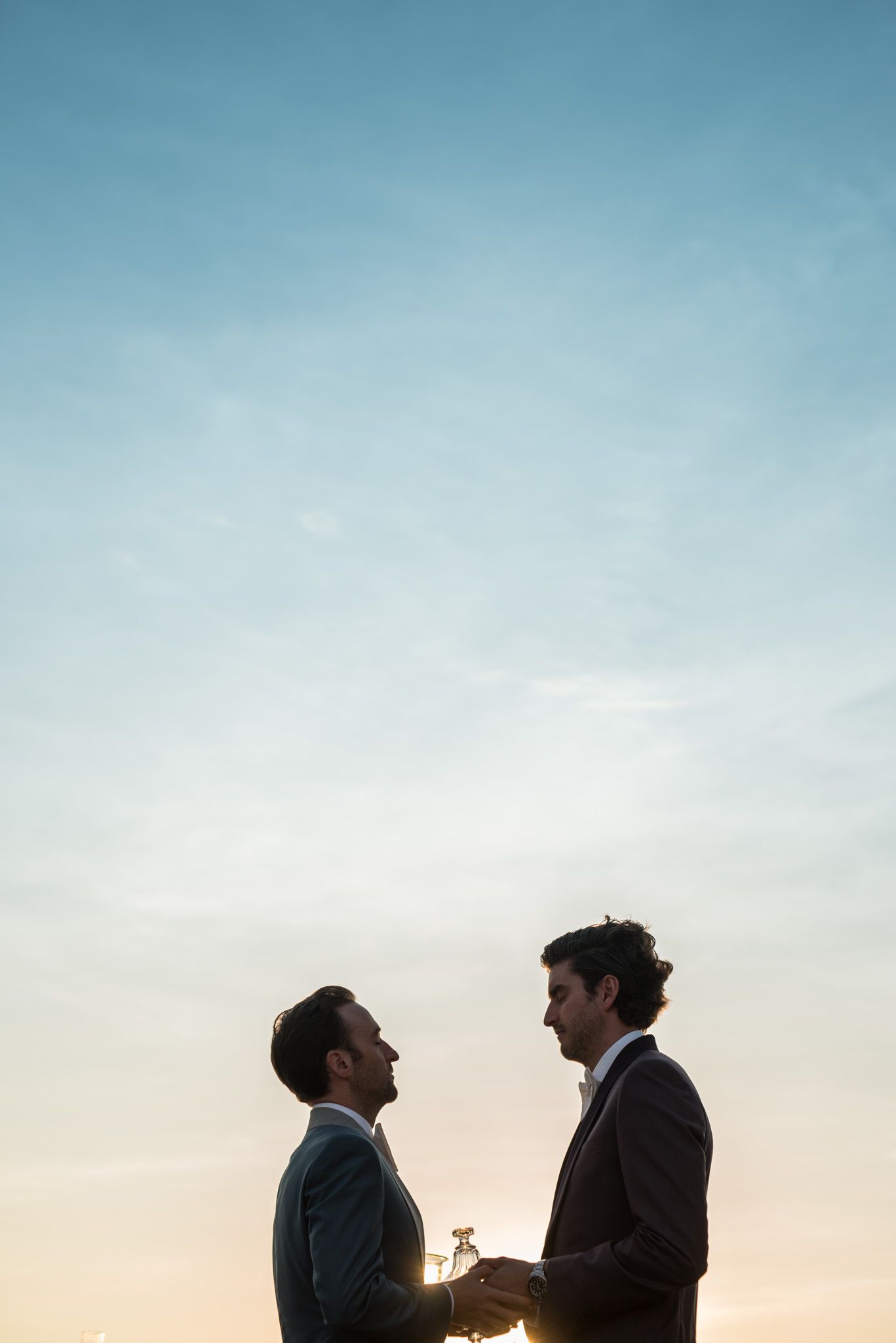 Two people in formal suits share an intimate moment against a sunset sky with soft blue gradient background.